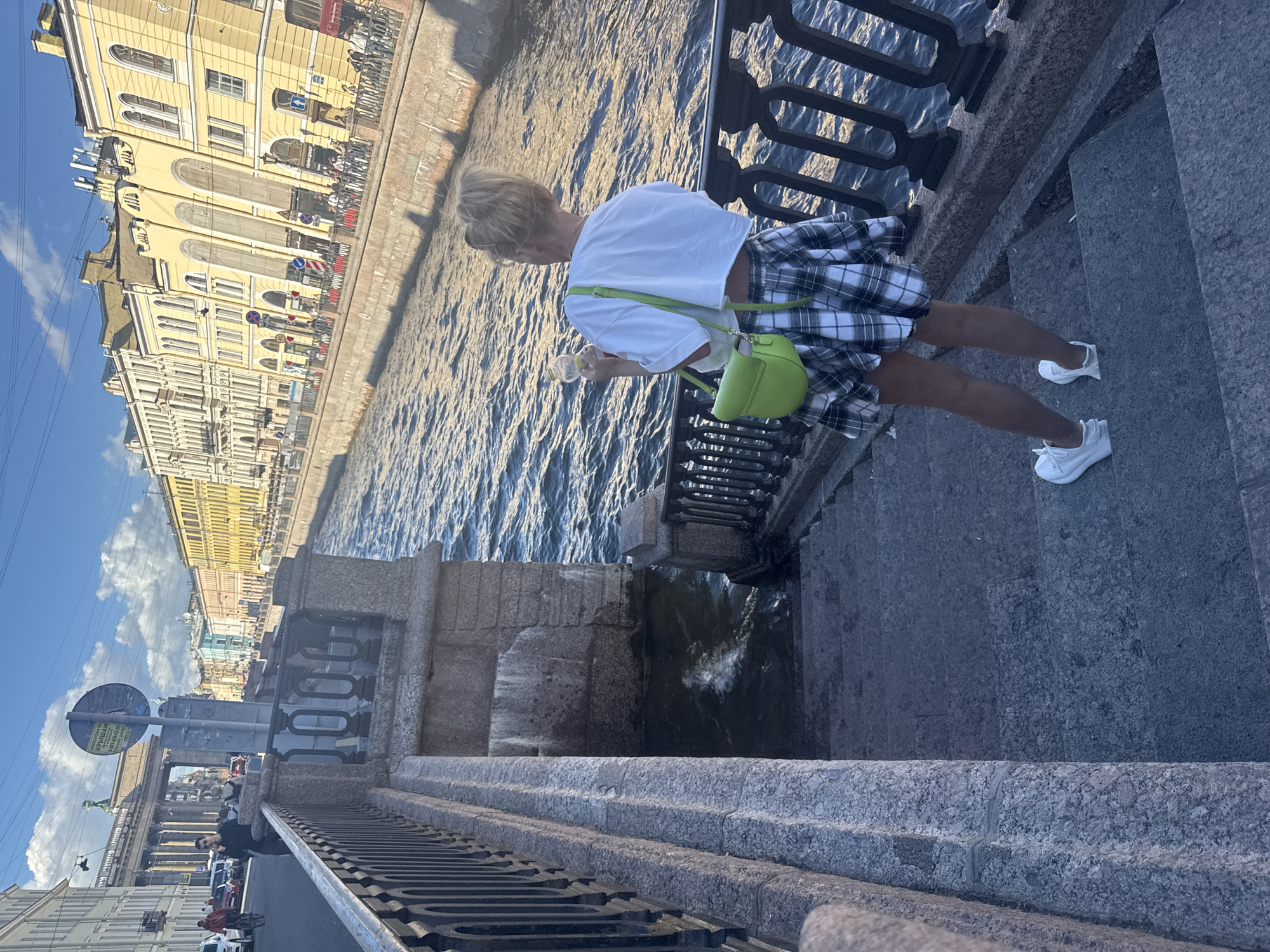 A woman is standing on a granite staircase looking out at a canal in Saint Petersburg, Russia. She has blonde hair pulled up in a bun and is wearing a white crop top, a plaid skirt, and white sneakers. A bright green purse is slung over her shoulder. The canal is lined with tall, colorful buildings, typical of Saint Petersburg's architecture. In the background, a large archway can be seen, likely a significant historical monument. The water in the canal is rippling, and the sky is blue with some white clouds. Several people are visible on the opposite bank of the canal, some walking and others on bicycles. The scene appears to be a bright, sunny day.