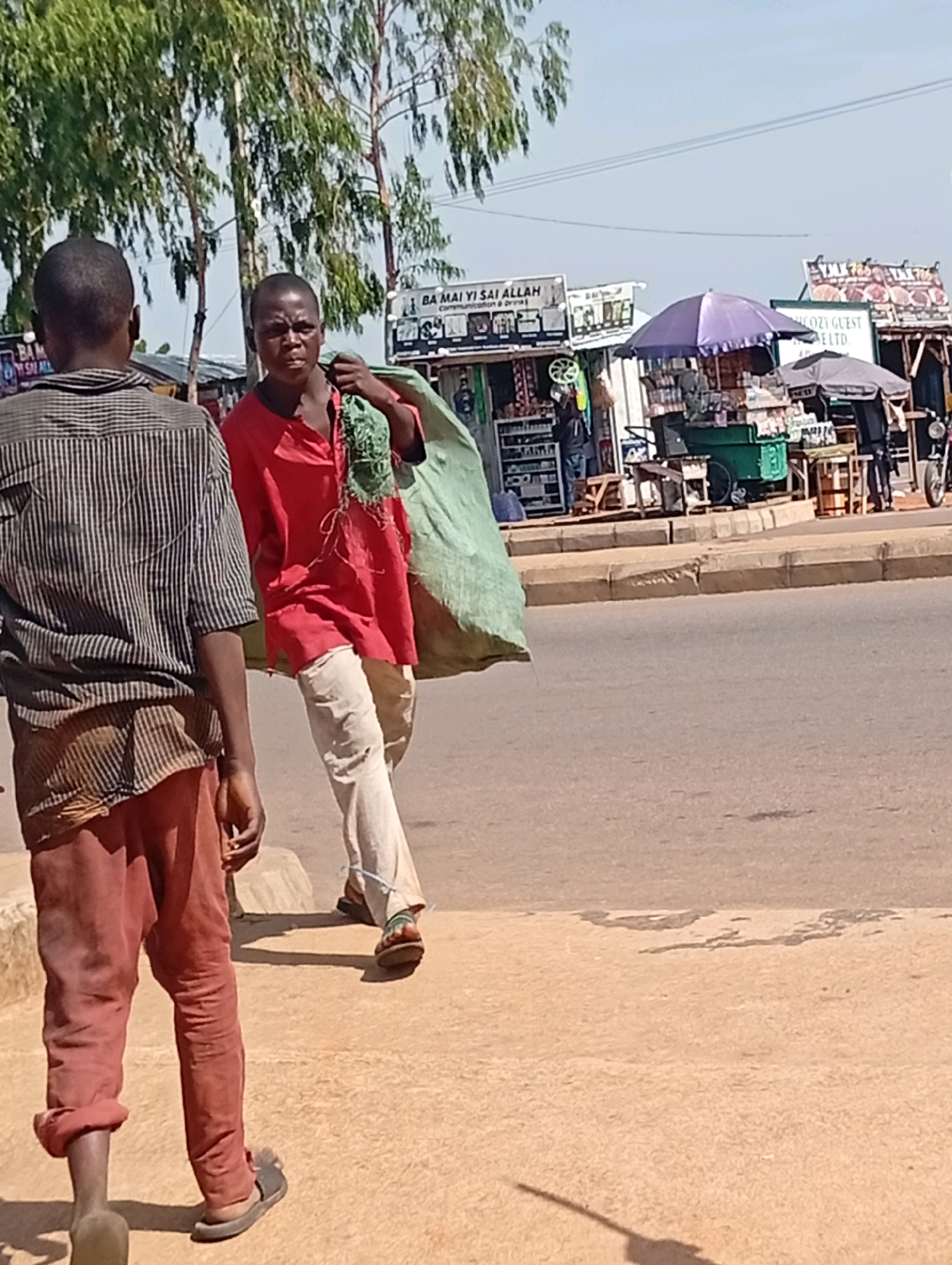 In Tumfure, Nigeria, two young males are depicted walking on a paved walkway next to a street. The individual on the left, viewed from the back, wears a striped shirt and reddish-brown pants. The person on the right faces forward, carrying a large, light green bundle over their shoulder. They are dressed in a red shirt and light-colored pants. Behind them, several market stalls are visible, featuring various goods and umbrellas, indicating a commercial area. The environment appears to be outdoors with trees and a clear sky. The ground is a mix of pavement and sandy soil.