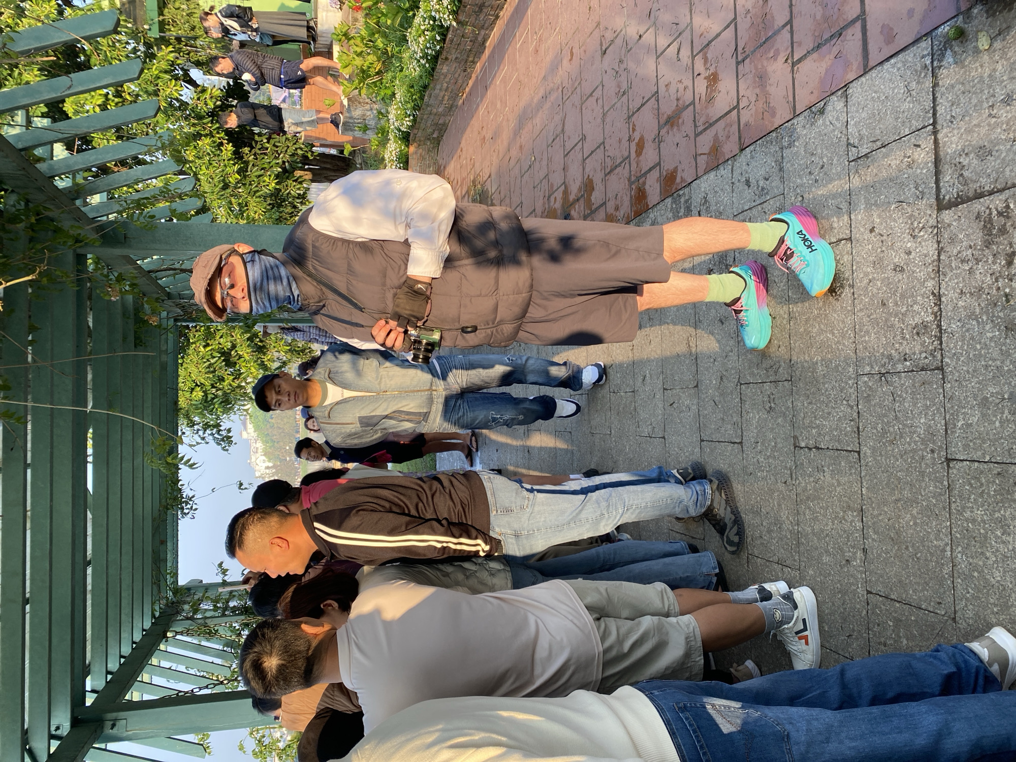 The image shows a group of people lined up in Da Lat, Vietnam, under a green wooden structure covered in foliage. The people are standing or leaning forward, seemingly looking at something. The person at the front of the line, facing the camera, is wearing shorts, a padded vest, a hat, a mask, and brightly colored Hoka shoes. They are holding a camera. Behind this person are a number of others, dressed in casual clothing such as jeans, shorts, t-shirts, and jackets. Some of them are wearing sneakers. The setting appears to be a public space with a stone-paved ground. In the background, other people and a building can be seen. The time of day appears to be afternoon, judging by the lighting.