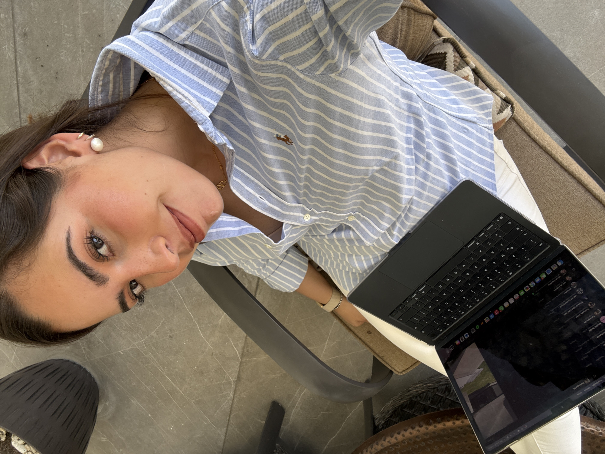 A woman sits with a laptop in an outdoor setting, likely in San Bernardino Tlaxcalancingo, Mexico. She wears a blue and white striped button-down shirt and white pants. A gold necklace with a pendant is visible at her neck. She has an earring in her left ear and is looking at the camera with a slight smile. A black laptop sits on her lap. The environment includes a chair, a cushion with a patterned design, and a floor with a slightly visible pattern.