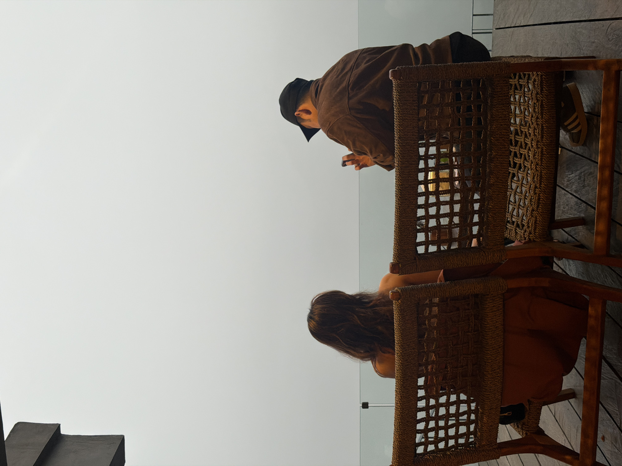 Two individuals are seated at a table on a wooden deck overlooking a hazy expanse. The person on the left, facing away from the camera, has long, dark, wavy hair and is wearing an orange dress. The person on the right, also facing away, is wearing a brown jacket and a black baseball cap and is looking at a mobile phone. The chairs are made of woven material and wood. A glass railing is visible in the background, separating the deck from the obscured view. The overall atmosphere suggests a cloudy or foggy day. The scene is located in Kintamani, Indonesia.