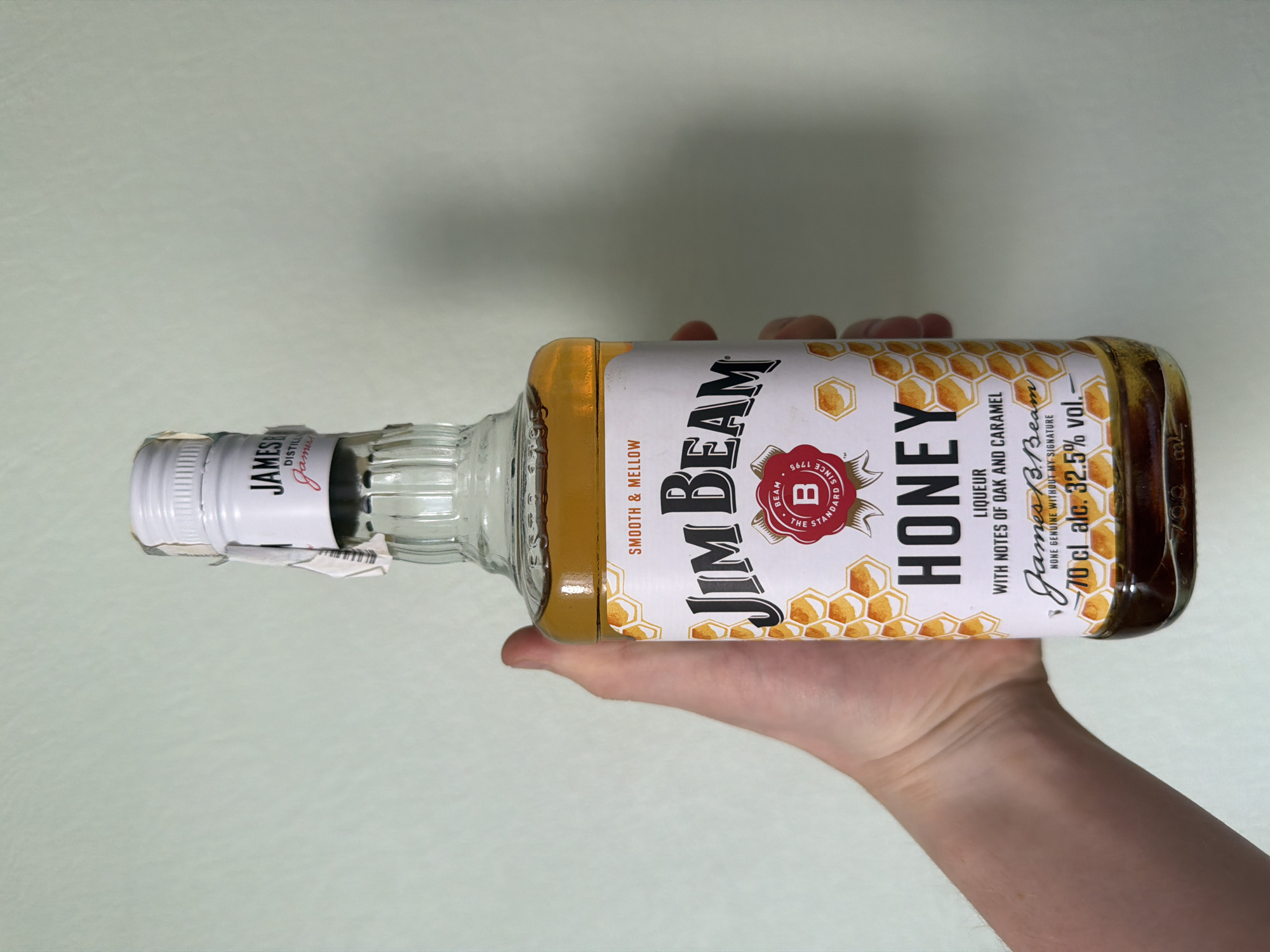 A person's hand is holding a bottle of Jim Beam Honey liqueur. The bottle is made of clear glass and is filled with a golden-colored liquid. The label on the bottle features a honeycomb pattern, the Jim Beam logo, and the words "HONEY," "LIQUEUR," and "WITH NOTES OF OAK AND CARAMEL." Below this, it says "James B. Beam" in script, followed by "NONE GENUINE WITHOUT MY SIGNATURE," and then "70 cl alc. 32.5% vol." The cap of the bottle is white and has a paper seal attached. The background is a plain, light green wall.