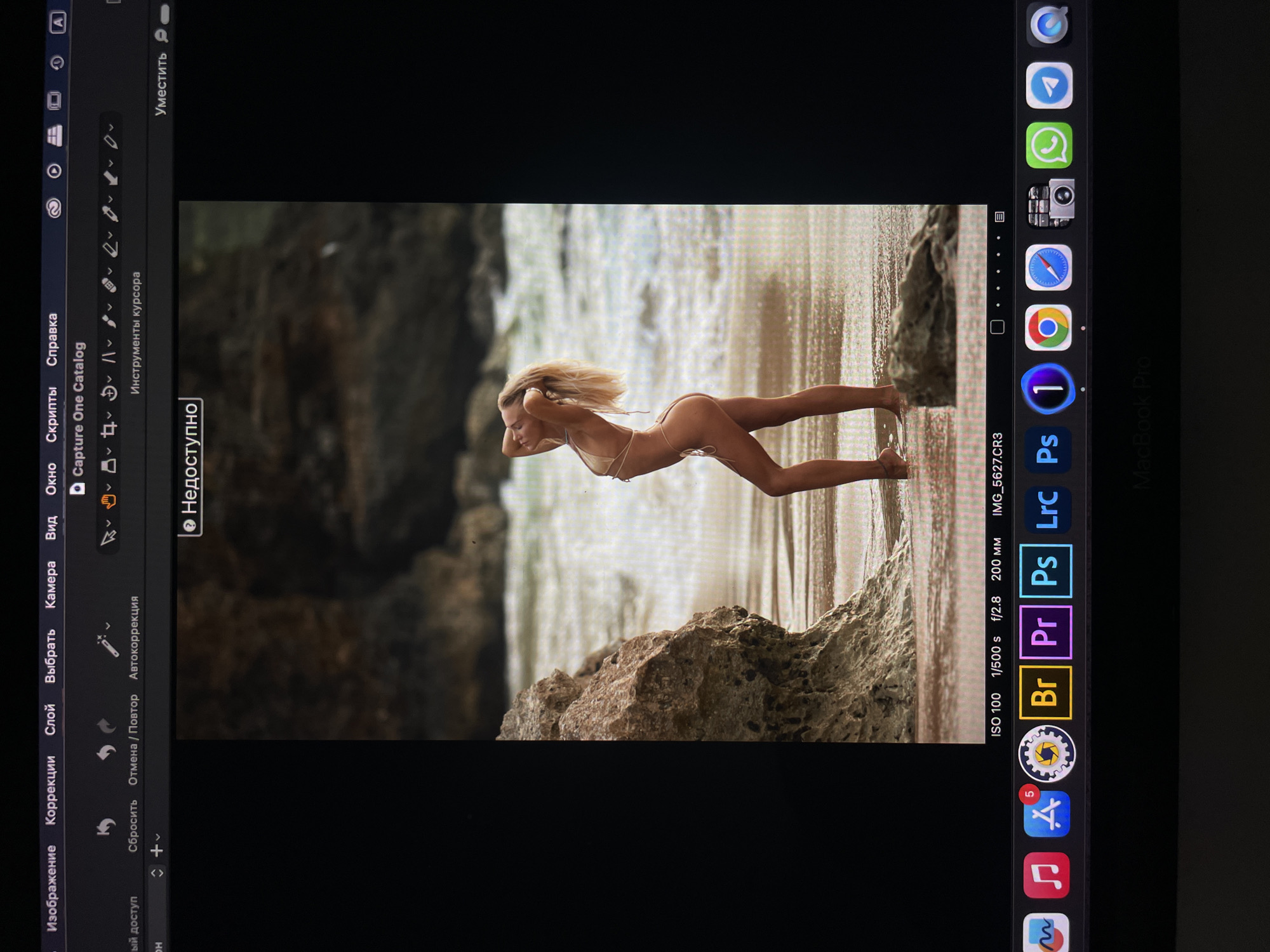 The image shows a woman in a bikini posing on a beach near rocks. The photograph is being edited on a MacBook Pro using Capture One software. The image metadata indicates it was shot at ISO 100, 1/500s, f/2.8, 200mm and the file is IMG_5627.CR3.