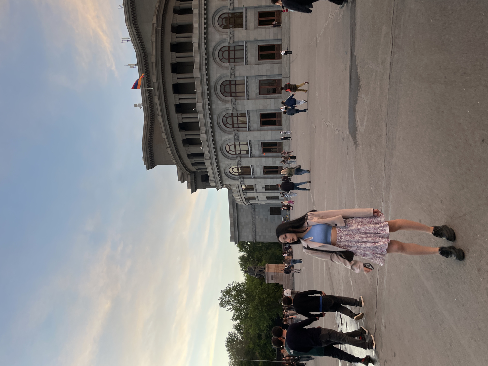 The image depicts a plaza in Yerevan, Armenia, likely Republic Square, recognizable by the distinctive semicircular government building. The scene is populated by many pedestrians. In the foreground, a young woman with dark hair is standing. She is wearing a light blue top, a floral skirt, and a beige jacket. She is standing on a paved area. In the background, a statue can be seen amidst some trees. An Armenian flag flies atop the government building. The sky is light blue with pale clouds.