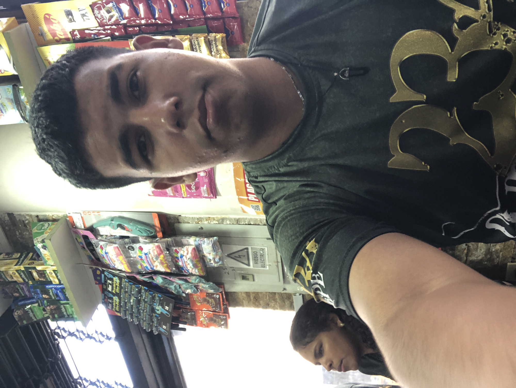A selfie appears to be taken in a small store in Juan Griego, Venezuela. A man with short dark hair and a grey shirt with a gold-colored design is in the foreground. In the background, a woman with dark hair and a dark shirt is partially visible. Behind the man, shelves display various food and household items, including snack foods like Adobo Completo, and other products. A sign that reads "RIESGO ELECTRICO" (Electrical Risk) is present on a wall.