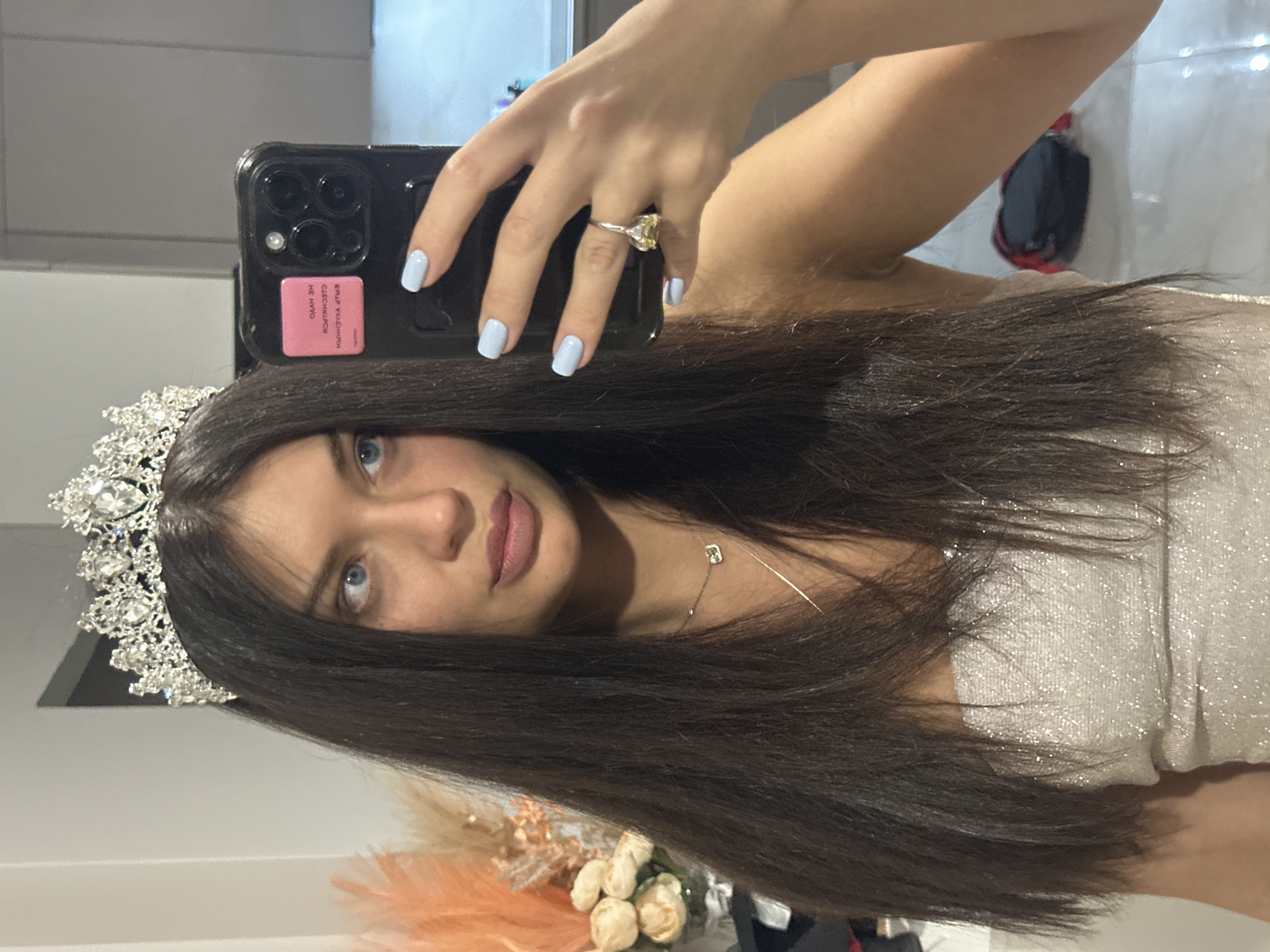 A young woman is depicted taking a mirror selfie, with the image captured in a portrait orientation, then rotated 90 degrees counter-clockwise. She has long, dark hair flowing over her shoulders, striking light blue eyes, and is wearing light pink lipstick. Adorning her head is a prominent silver-toned tiara or crown embellished with sparkling clear stones. She is dressed in a shimmery, light-colored (possibly silver or white) strapless garment, suggesting formal wear.
Her left hand is visible, holding a black smartphone (likely an iPhone given the camera array), which partially obscures her face. Her fingernails are neatly painted with a light blue polish. On her left ring finger, she wears a distinctive ring featuring a large yellow gemstone. A delicate silver necklace with a small square pendant hangs around her neck.
The background, visible through the mirror's reflection, shows interior elements of a room, including what appears to be a light-colored wall and some indistinct objects. In the lower right portion of the frame, artificial light-colored flowers and some orange or peach feathery decorative items are arranged on a surface.
A pink rectangular sticker is affixed to the back of her phone case, displaying inverted white text. The script is difficult to decipher clearly due to the reflection and angle, but it appears to be in a non-English language, possibly Cyrillic. The overall scene suggests the woman is dressed up, perhaps for a special event, and is capturing her reflection.