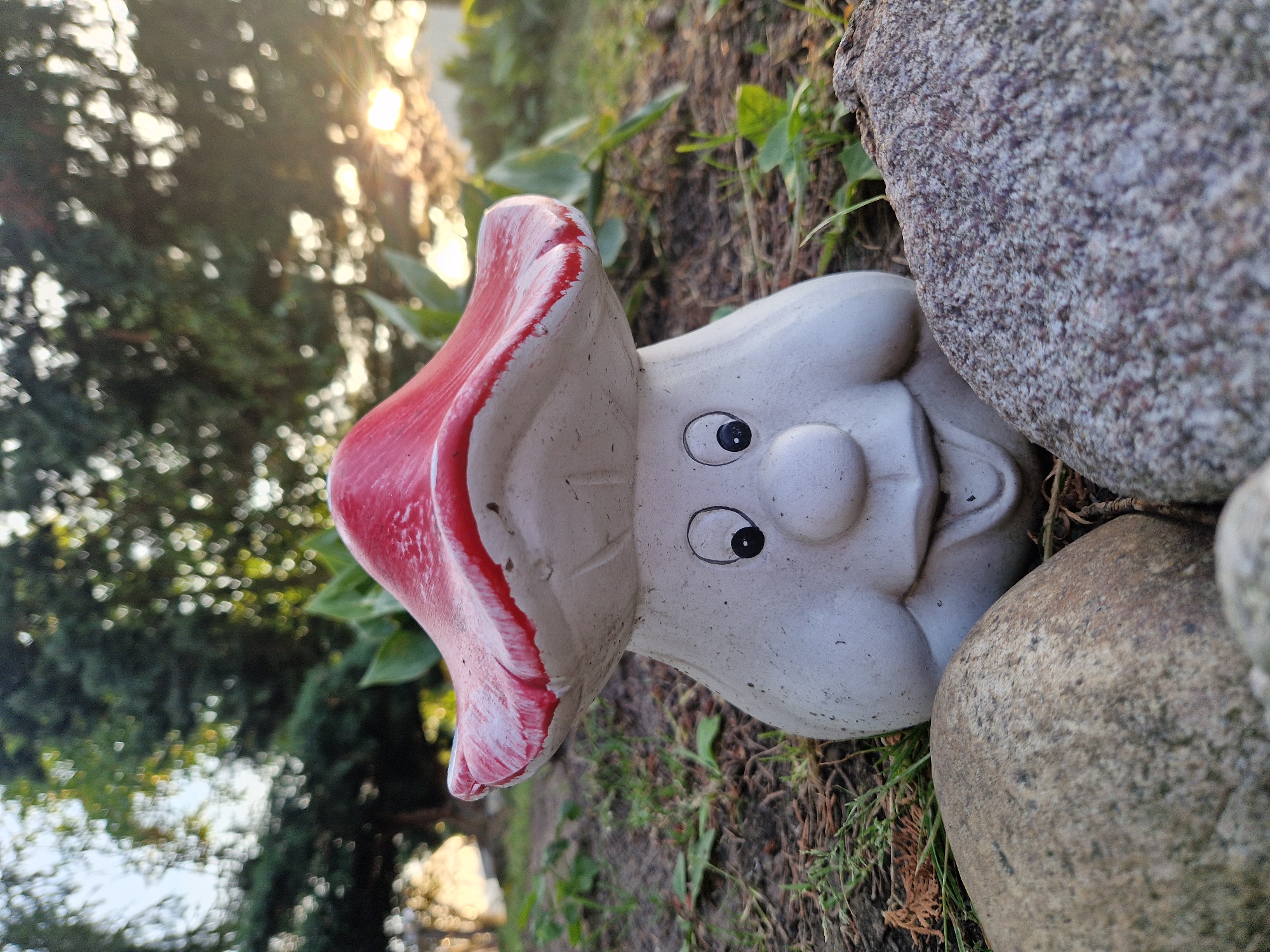A low-angle, medium shot captures a ceramic garden gnome shaped like a mushroom, sitting on the ground outdoors. The gnome has a white, bulbous body with a smiling face, featuring black circular eyes and a round nose. Its mushroom cap is red with white highlights along the rim. Two large, textured rocks are positioned in the foreground, one on the left and one on the right, partially obscuring the gnome's base. The background shows blurred greenery, likely trees and bushes, with sunlight filtering through the foliage, creating a bokeh effect of heart shapes. The scene appears to be set in a garden or natural outdoor area.