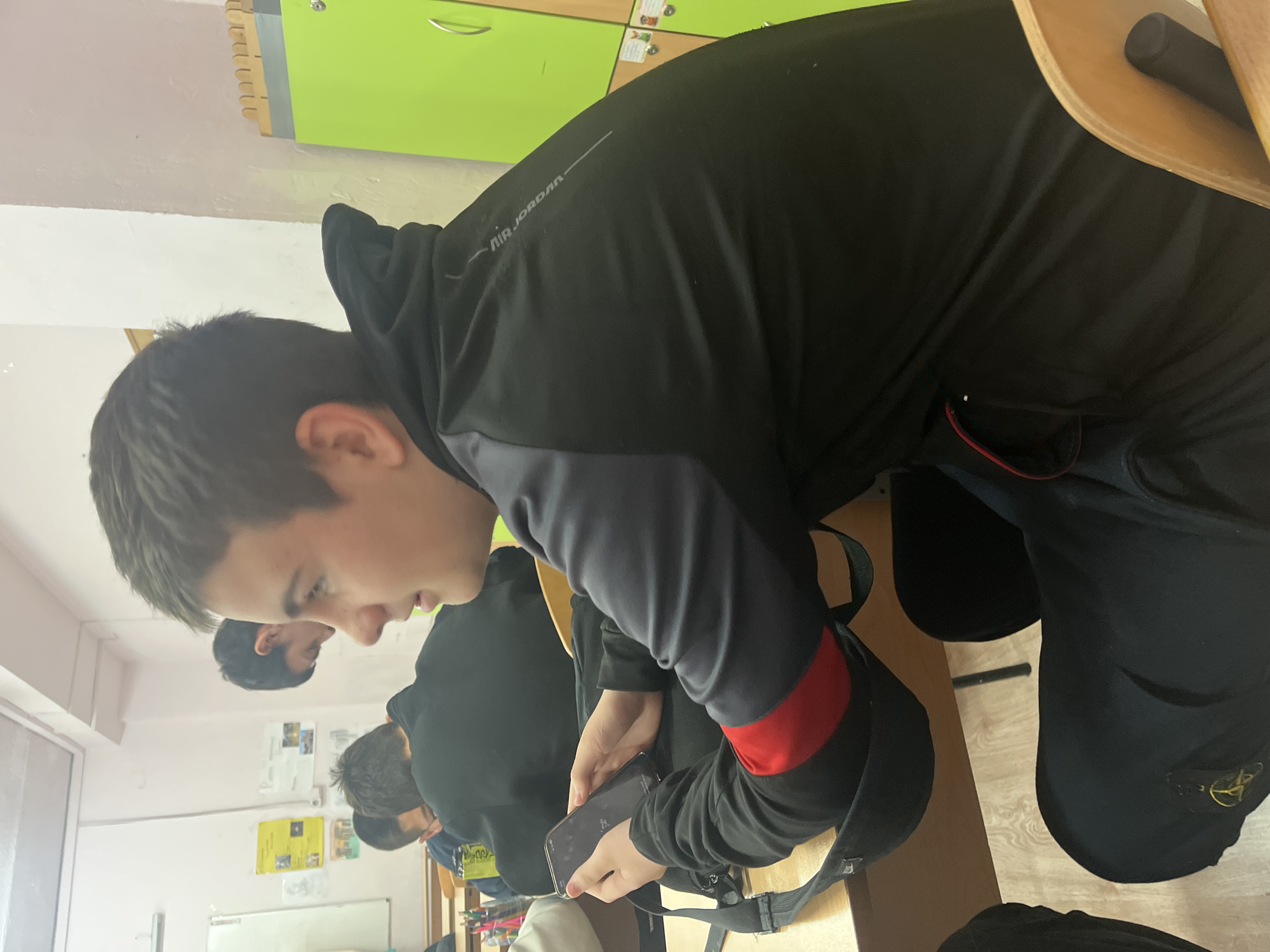 A classroom setting in Sofia, Bulgaria shows a group of students. One boy, wearing a black Air Jordan hoodie with a red band on the sleeve, is looking at a smartphone. There are other students seated in the background and educational materials posted on the wall. The classroom is equipped with lockers.