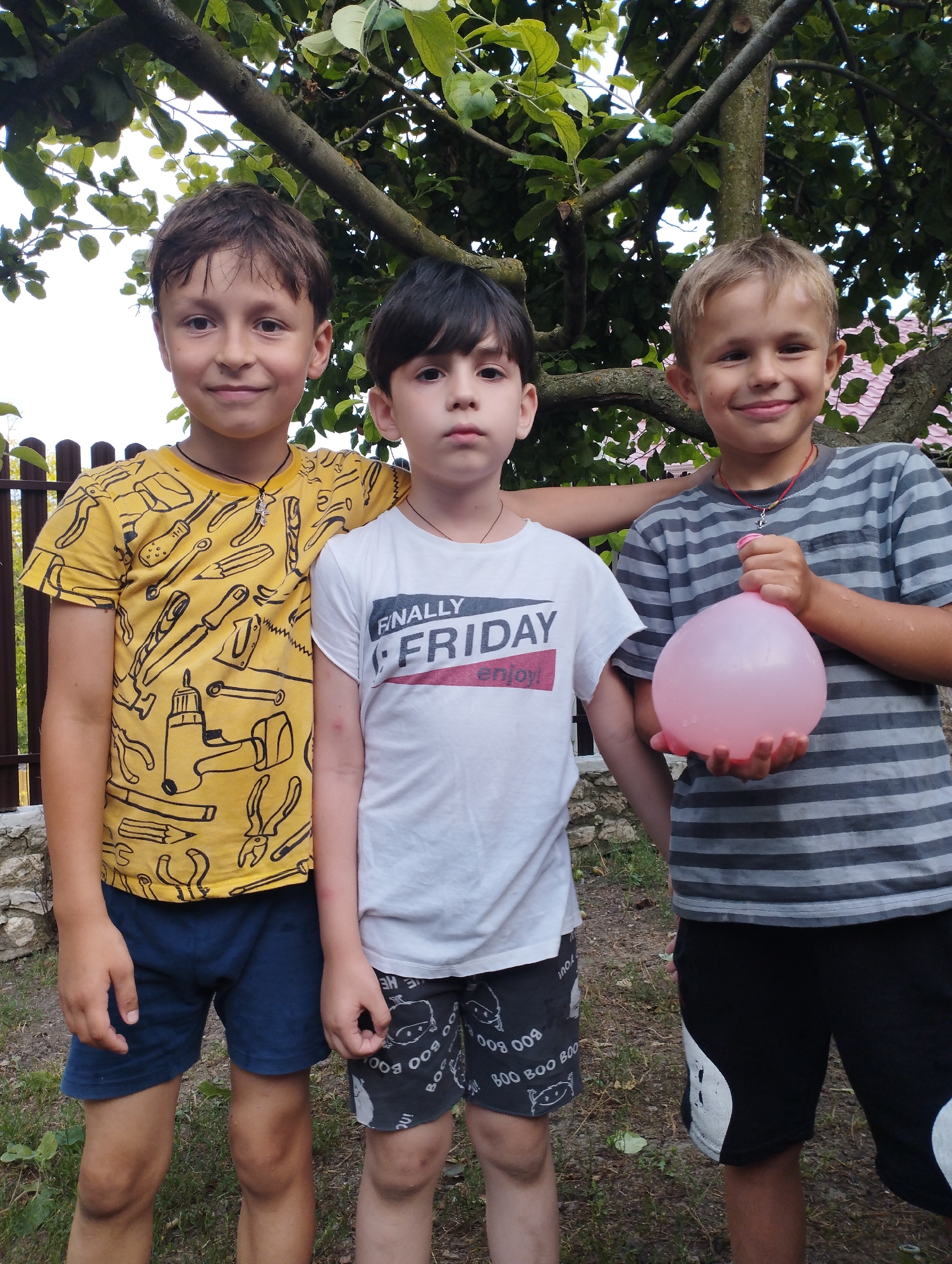 Three young boys stand together outdoors, likely in a backyard, under a tree with dense green foliage. The boy on the left wears a yellow t-shirt with a black tool print and dark blue shorts. The middle boy is dressed in a white t-shirt with "FINALLY FRIDAY enjoy!" printed on it, and dark shorts with a "BOO BOO" pattern. The boy on the right sports a grey and black striped t-shirt and black shorts. He holds a pink water balloon in his hands, suggesting an outdoor activity like a water fight. The overall scene appears casual and joyful, possibly during a warm, sunny day, as indicated by the natural lighting and the children's attire. The background includes a wooden fence and some stone work, hinting at a residential setting in Fălești, Moldova. The boys are all smiling, conveying a sense of fun and camaraderie.