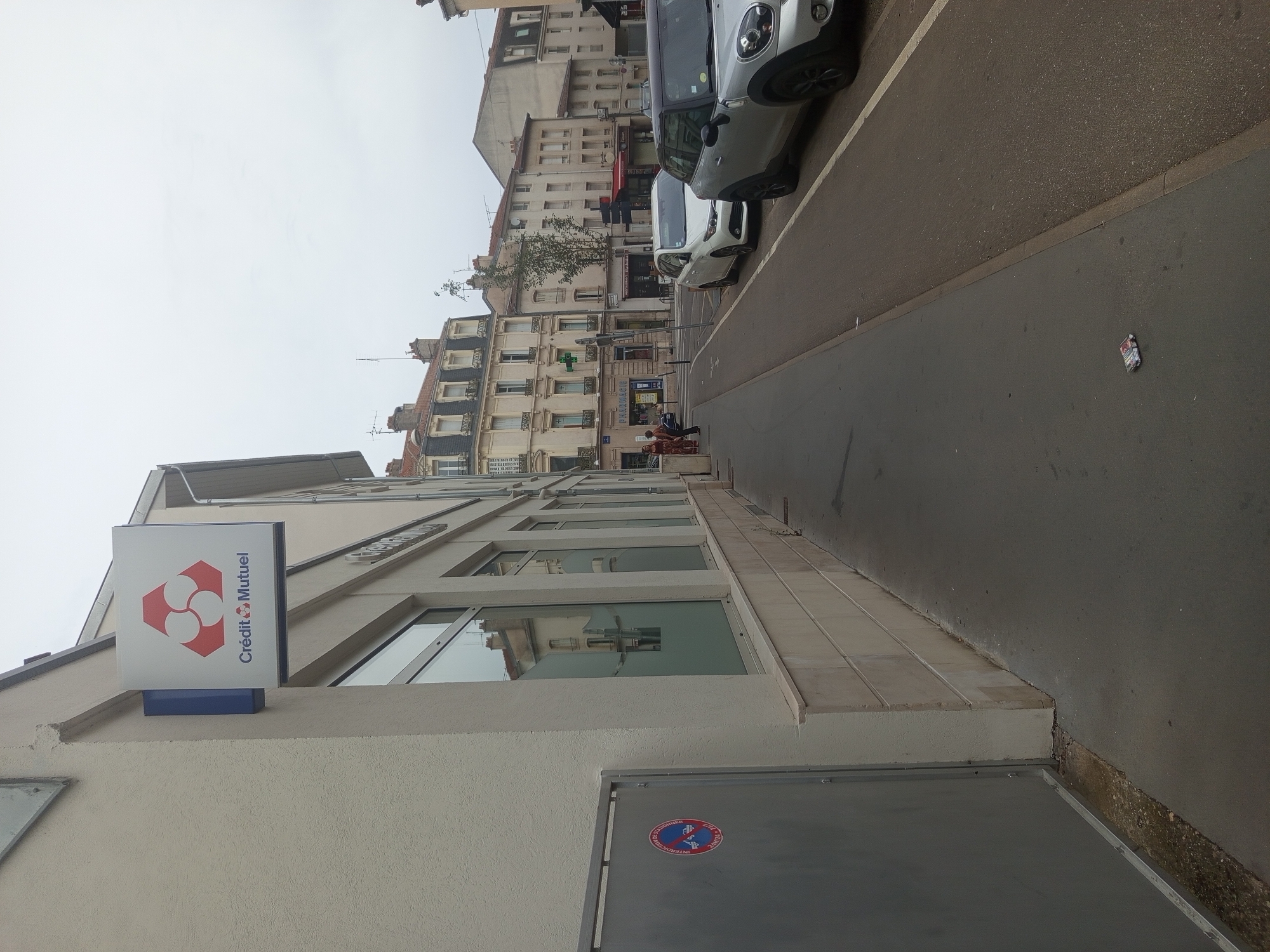 The image depicts a street scene in Nancy, France, specifically on Rue de la République. On the left, a light-colored building displays a sign for "Crédit Mutuel," a French bank, featuring its red and white circular logo. Adjacent to the building is a grey metal enclosure with a circular "No Parking" sign. The street itself is paved, with a sidewalk running alongside the buildings. Several cars are parked along the street, including a silver Mini Cooper and a white hatchback. In the background, a row of multi-story buildings with traditional European architecture lines the street. Two individuals are visible walking on the sidewalk in the distance. The sky is overcast, suggesting a cloudy day.