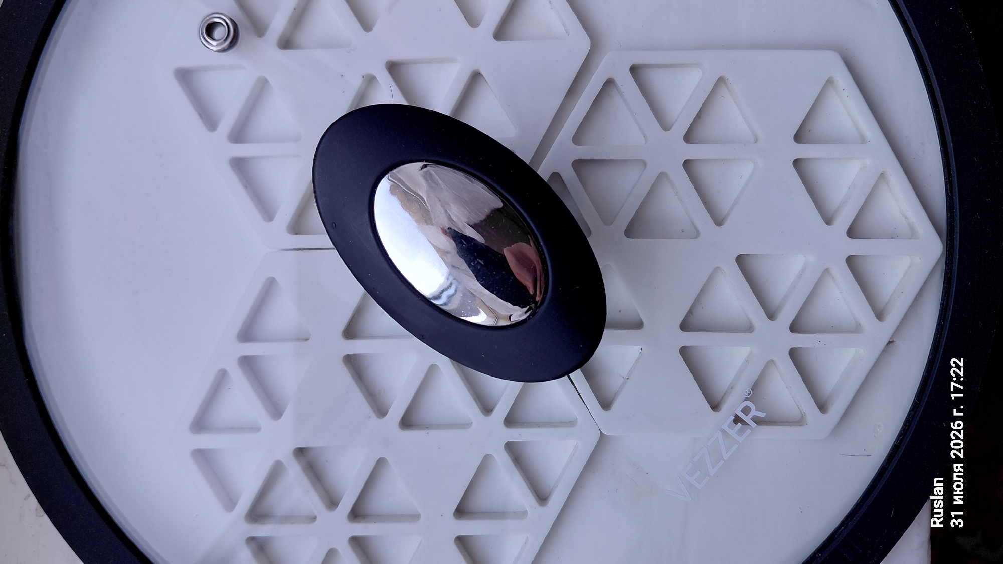 The image shows a close-up, top-down view of a white lid with a black rim. The lid has a pattern of raised hexagonal shapes, each divided into six triangles. In the center of the lid is a black, oval-shaped handle with a shiny, metallic knob. The text "VEZZER" is visible on the lower left side of the lid, partially obscured by the geometric pattern. In the bottom left corner of the image, text reads "Ruslan 31 июля 2026 г. 17:22". This implies the image was taken by someone named Ruslan on July 31, 2026, at 5:22 PM. The location of Oleksandriia, Ukraine is not depicted or suggested by any elements in the image.
