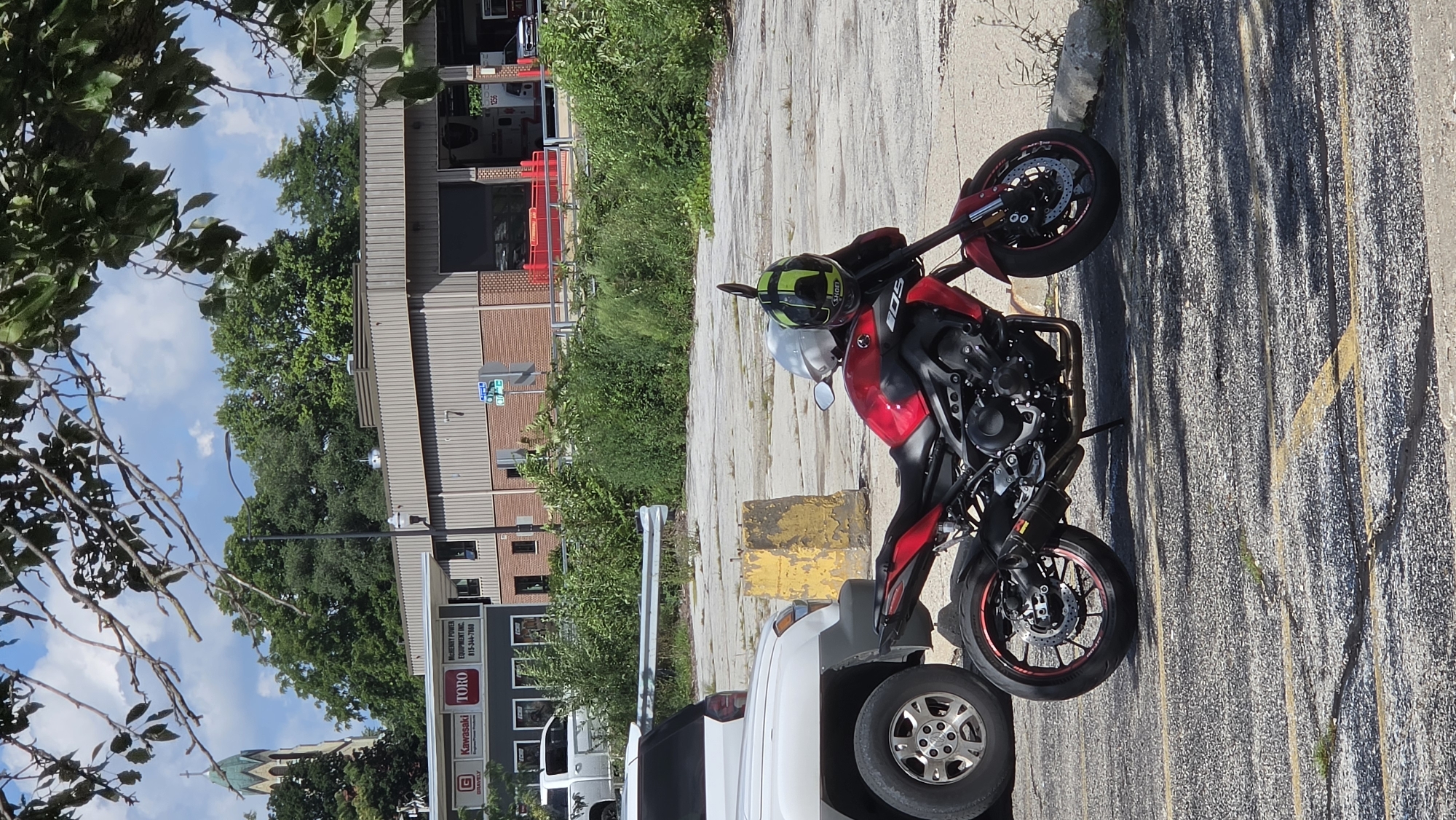 In McHenry, Illinois, a red Yamaha motorcycle is parked in an asphalt lot, with two helmets resting on its handlebars. One helmet is white, and the other is black and lime green. To the left of the motorcycle, a white SUV is parked. In the background, a brick building with a gray facade features several large garage doors, some of which are open. Signs on the building indicate it is a Kawasaki and Toro dealer, with a phone number listed as 815-344-7000. A steeple belonging to a church is visible above the building, and trees and foliage surround the area. The sky is partly cloudy.