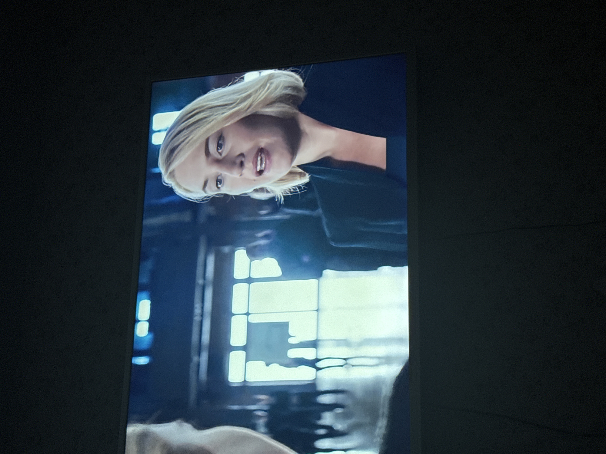 The image shows a screen displaying a woman with blonde hair looking up and to the right. She has fair skin, blue eyes, and appears to be speaking. She is wearing a dark top. Behind her is a blurred background, including a window with multiple panes. The surrounding area is dark, suggesting the image is being viewed in a dimly lit room. There is no information from the image to suggest that the image was taken in Mytishchi, Russia.
