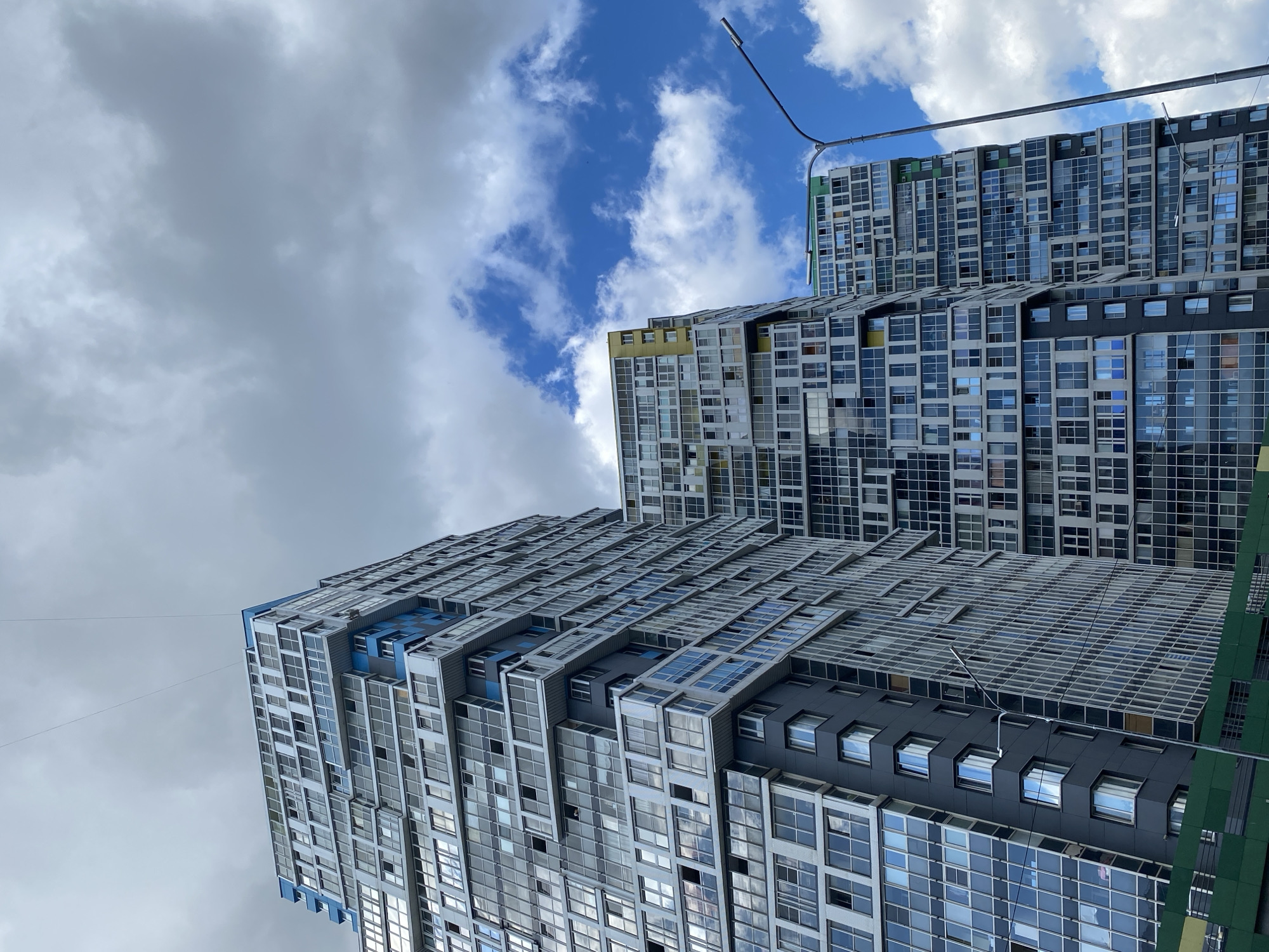 This media file features a low-angle shot of modern apartment buildings under a cloudy sky. The buildings are tall and constructed with a mix of glass, concrete, and blue accents. The architecture is characterized by repetitive window patterns and balconies, giving a sense of a densely populated urban environment. In the background, a street lamp extends into the frame, suggesting an outdoor setting. The sky is a mix of dramatic grey clouds and patches of bright blue, indicating variable weather, possibly a partly cloudy day. No people are visible in the frame. The scene appears to be set in Kudrovo, Russia, as per the provided context. The overall impression is one of contemporary urban development and architecture.