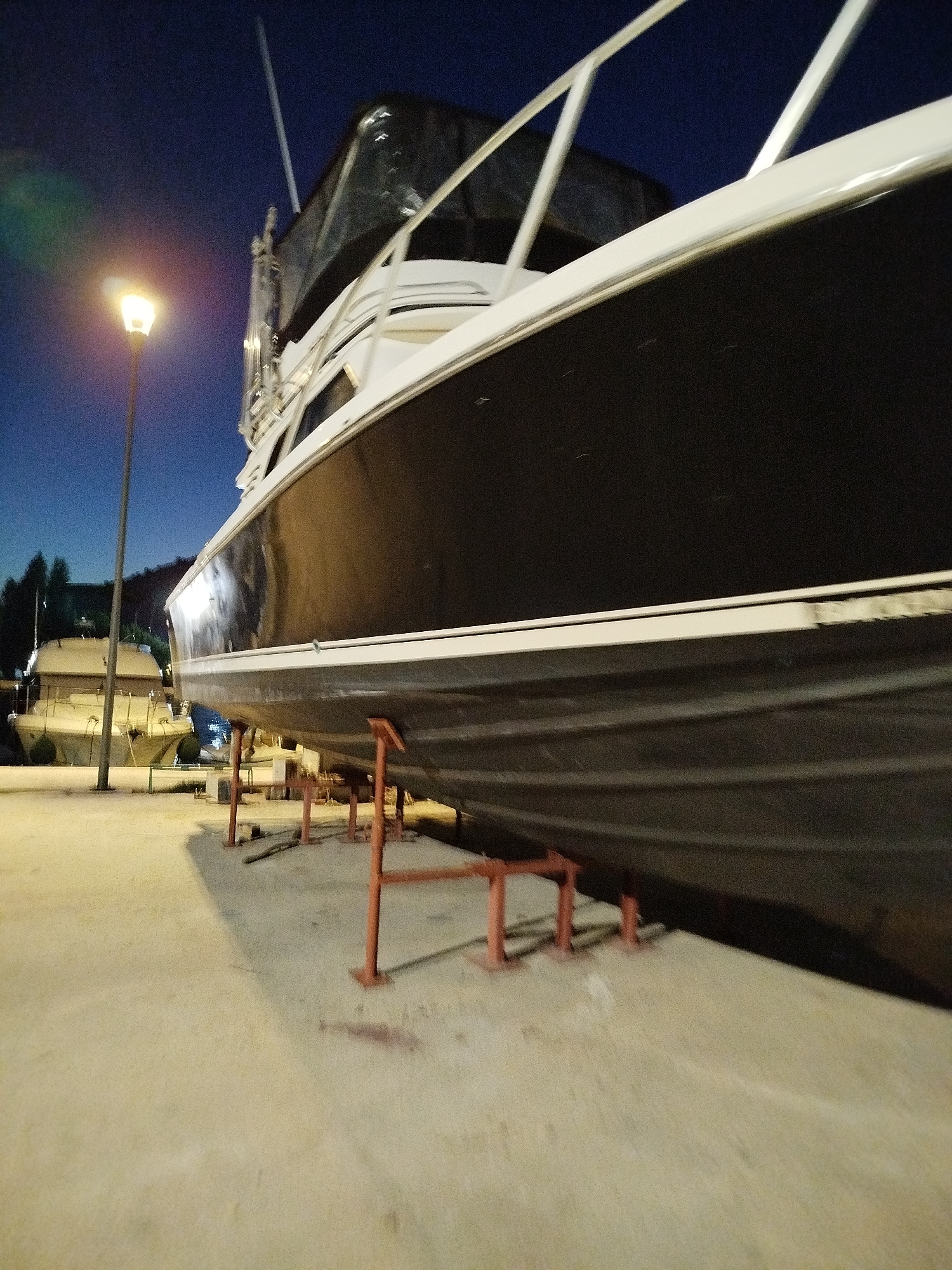 The image depicts a nighttime scene at a harbor in Omiš, Croatia. A large, dark-hulled boat rests on red stands on a concrete surface. The boat is angled towards the viewer, with its white upper structure and railings visible. In the background, another boat is partially visible, also resting on stands. A street lamp with a warm light illuminates the foreground, casting shadows. The sky is dark blue, indicating it is night. The environment appears to be a marina or shipyard, with the concrete pier and the presence of multiple boats on stands suggesting maintenance or storage. There are no people visible in the image.
