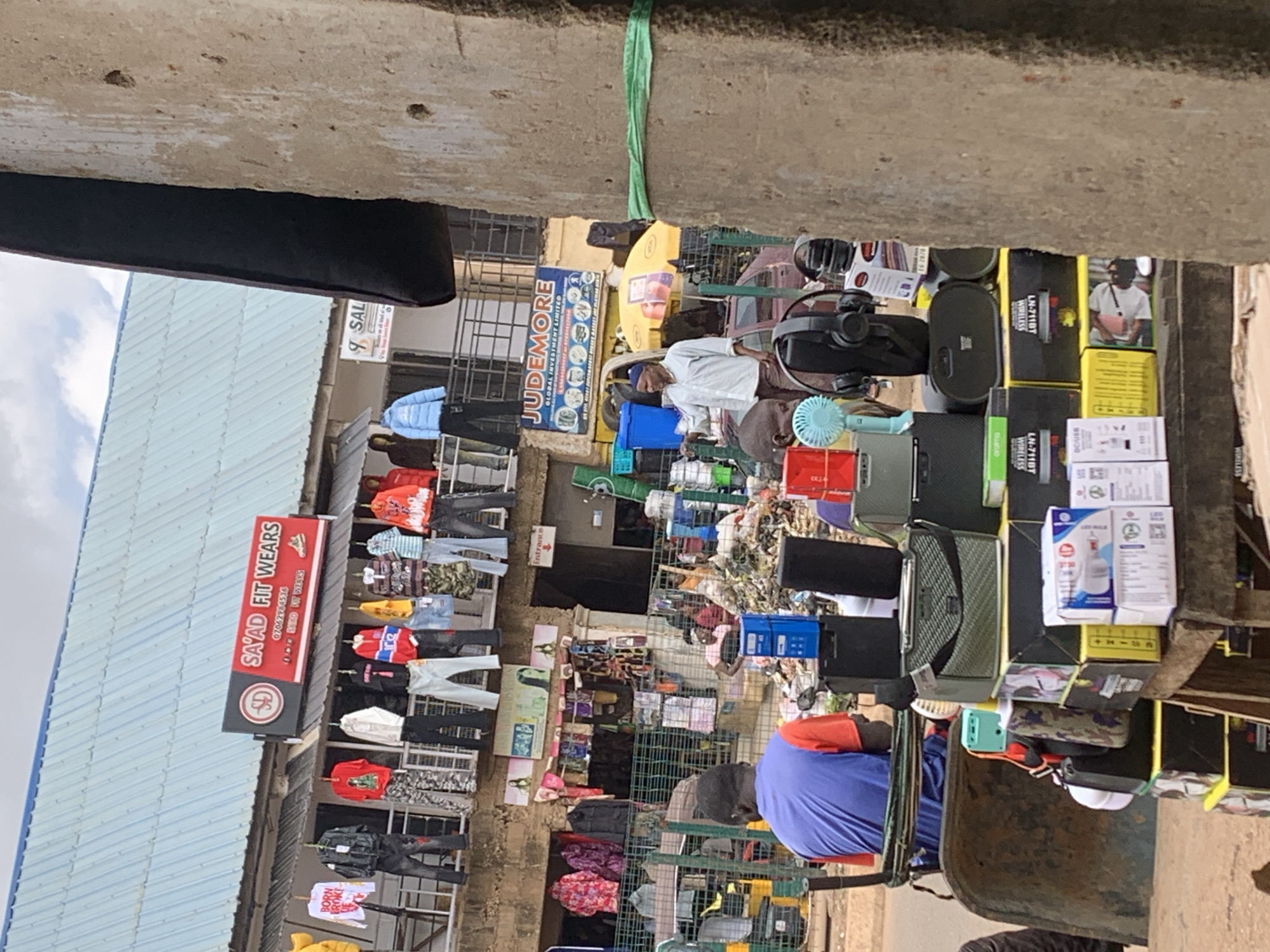 The image depicts a bustling market scene in Bukuru, Nigeria. It shows a collection of stalls selling various goods, including clothing, electronics, and other merchandise. Several people are visible, either tending to the stalls or browsing the items. One stall prominently displays "SAYAD FIT WEARS," indicating a clothing vendor. Another stall has a sign for "JUDEMORE GLOBAL INVESTMENT LIMITED." Various electronic items, such as headphones and what appear to be phone accessories, are also displayed. The setting appears to be an outdoor market with a corrugated metal roof overhead and a rough concrete structure at the top of the image. The lighting suggests it is daytime. The overall atmosphere is one of commerce and everyday life in a Nigerian market.
