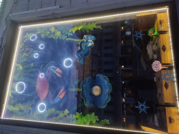 The image depicts a brightly lit window display of an aquarium or nautical-themed scene. On the left side, there's a painted scene of a crab and other marine life surrounded by illuminated circles. The right side of the display features a painted scene of an underwater themed window with a large pearl and a sea horse. The reflection in the window reveals an urban scene at night, presumably in Moscow, Russia, given the architecture.