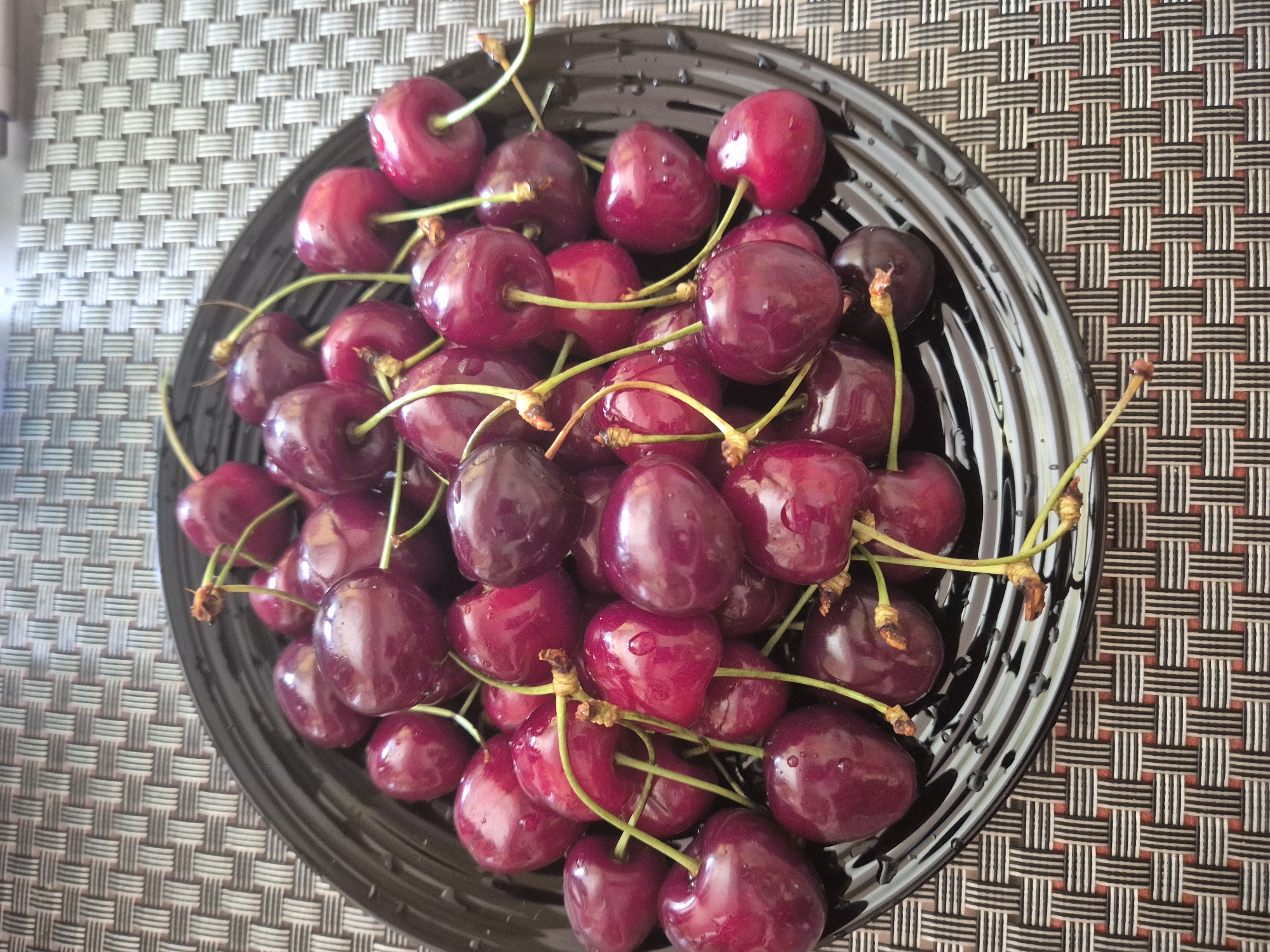 A close-up, overhead shot shows a black bowl filled with dark red cherries. The cherries are wet, with water droplets visible on their surfaces. Most of the cherries have stems attached, which are light green in color and some have small brown stems at the base where they connect to the fruit. The bowl itself is dark and has a glossy finish, reflecting the light in thin, curved lines. The bowl is placed on a textured placemat that has a woven pattern of alternating white, black, and thin red vertical lines. The lighting is bright and even, suggesting it could be daytime, though no specific location cues are evident. The image focuses solely on the bowl of cherries and the placemat, with no other objects or people visible.