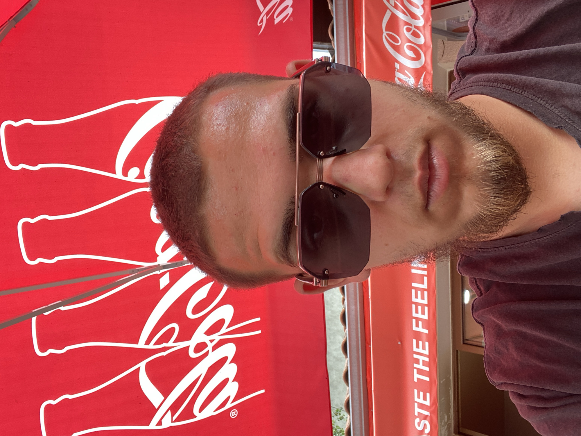 The image is a close-up, selfie-style shot of a young adult male wearing sunglasses and a dark t-shirt. He has short, light brown hair and a goatee. He is positioned in front of a red background with white Coca-Cola logos and bottle silhouettes. A red awning with white lettering that reads "TASTE THE FEELING" is visible below him. The setting appears to be an outdoor area, possibly a cafe or refreshment stand, given the awning and the visible building structure. The backdrop suggests it is daytime, and the lighting is bright. While the image contains visual elements associated with the city of Sevlievo, Bulgaria, through the recognizable Coca-Cola branding often found in commercial establishments globally, no definitive or specific markers within the image itself pinpoint the exact location as Sevlievo.