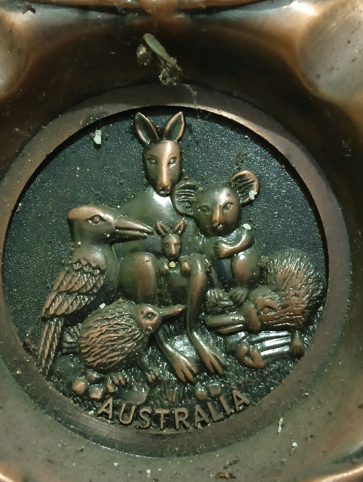 The image shows a circular, bronze-colored relief depicting several Australian animals. The animals include a kangaroo, a joey in its pouch, a koala, a kiwi bird, a kookaburra, and a echidna. The word "AUSTRALIA" is embossed in capital letters below the animals. The relief is set within a larger, darker circular area, which is part of a tarnished bronze bowl or ashtray. The surface of the bowl and the relief are covered in dust and debris, suggesting age and lack of recent cleaning. There are no people visible in the image. The scene is entirely composed of the relief and its immediate surroundings within the bowl. The location in the city of Sukosari, Indonesia, is not depicted or suggested in the image itself.