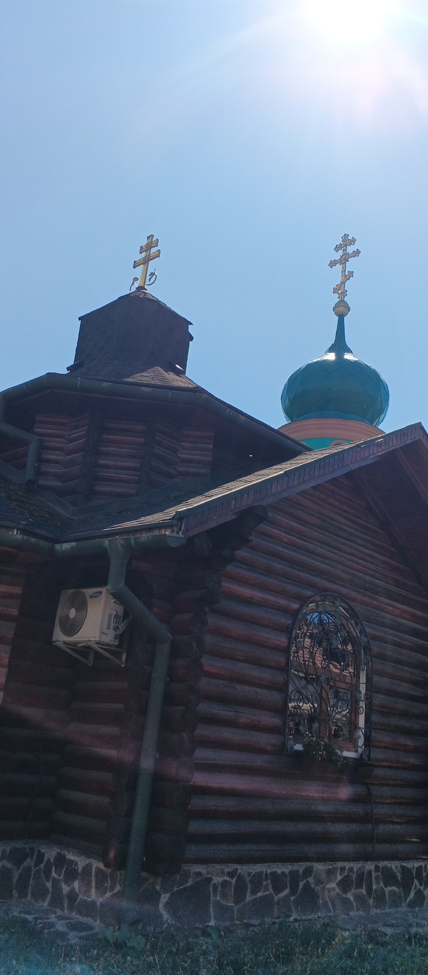 This is a low-angle, full-frame, outdoor, eye-level shot of a wooden Orthodox church, likely located in or near Mukachevo, Ukraine. The church features a prominent, dark-stained timber construction. Two crosses are visible atop the church structures. The larger cross, gold-colored, stands on a multi-tiered roof section, while a second, more elaborate gold cross tops a green, rounded dome. A decorative, wrought-iron window grille is visible on the side of the building, framing what appears to be stained glass. An air conditioning unit is mounted on the wooden wall, below a section of guttering. The lower portion of the church is constructed with rough, grey stone. The background is a clear, bright blue sky with a sun flare visible in the upper right corner. The foreground shows sparse green grass. The image is sharp and well-lit, suggesting it was taken on a sunny day.