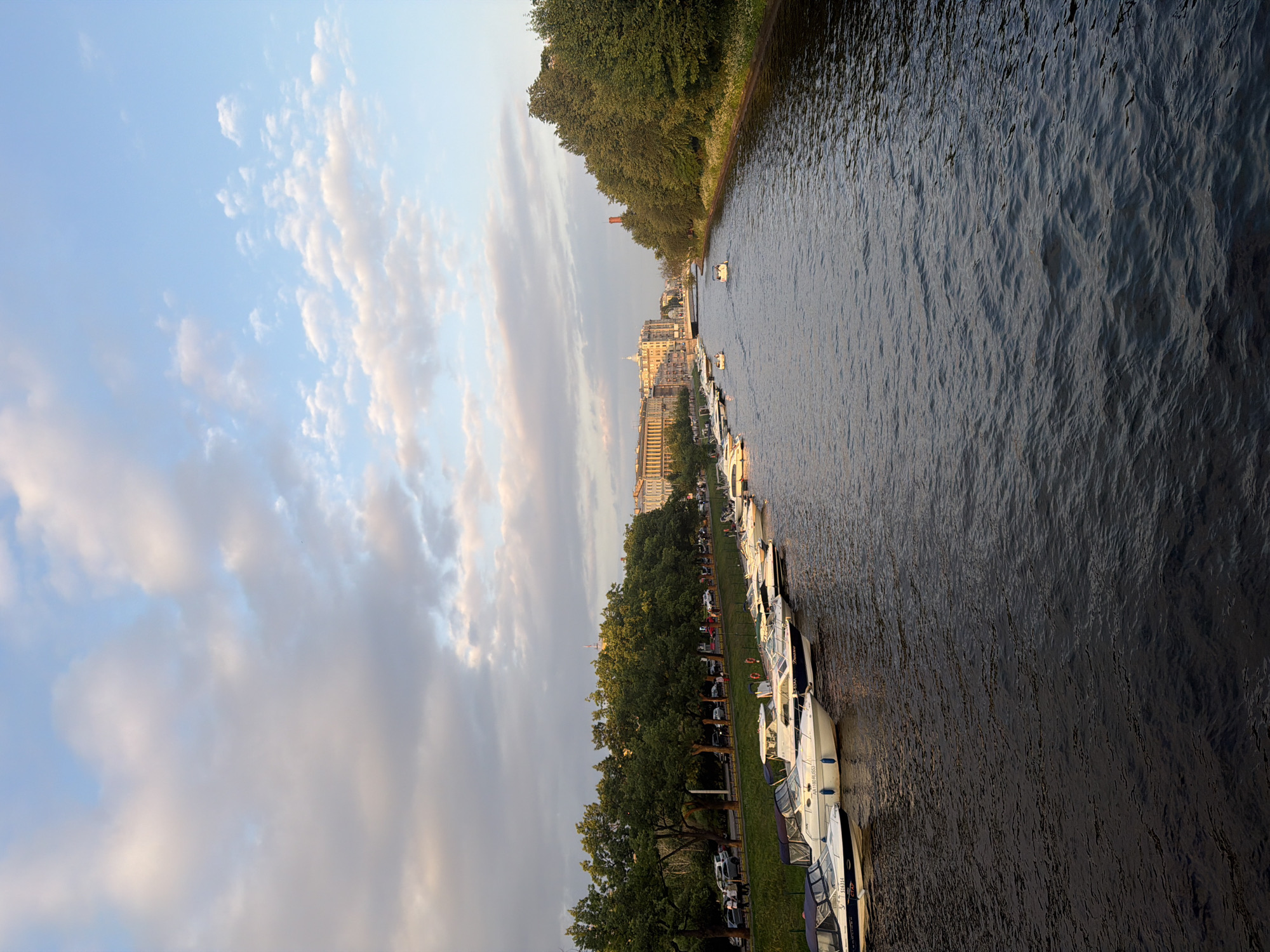 The image depicts a serene waterside scene, likely in the late afternoon or early evening, given the soft lighting. The sky is a gentle blue, adorned with wispy, elongated clouds that catch the warm hues of the setting sun. To the right, a body of dark, rippling water fills the frame, its surface reflecting the sky and surrounding elements.
Along the left bank of the water, a line of moored boats is visible, their white hulls contrasting with the dark water. The boats appear to be leisure craft, with some displaying names like "Diadora" and "KATHARINE ROSSI" on their sides. Beyond the boats, a lush green strip of grass is visible, bordered by a dense line of mature trees. The trees are full of leaves, suggesting a warmer season. Further inland, a cityscape emerges, with buildings of varying heights and architectural styles, bathed in a warm, golden light. Some structures appear to be older, with classic facades, while others are more modern.
The overall mood of the image is peaceful and tranquil. The calm water, the soft light, and the lush greenery create a sense of quiet beauty. There are no visible people in the foreground, adding to the feeling of stillness. The scene suggests a popular waterfront area, perhaps a marina or a scenic promenade, where people might gather to enjoy the evening. The presence of the boats indicates a setting where water-based activities are common.