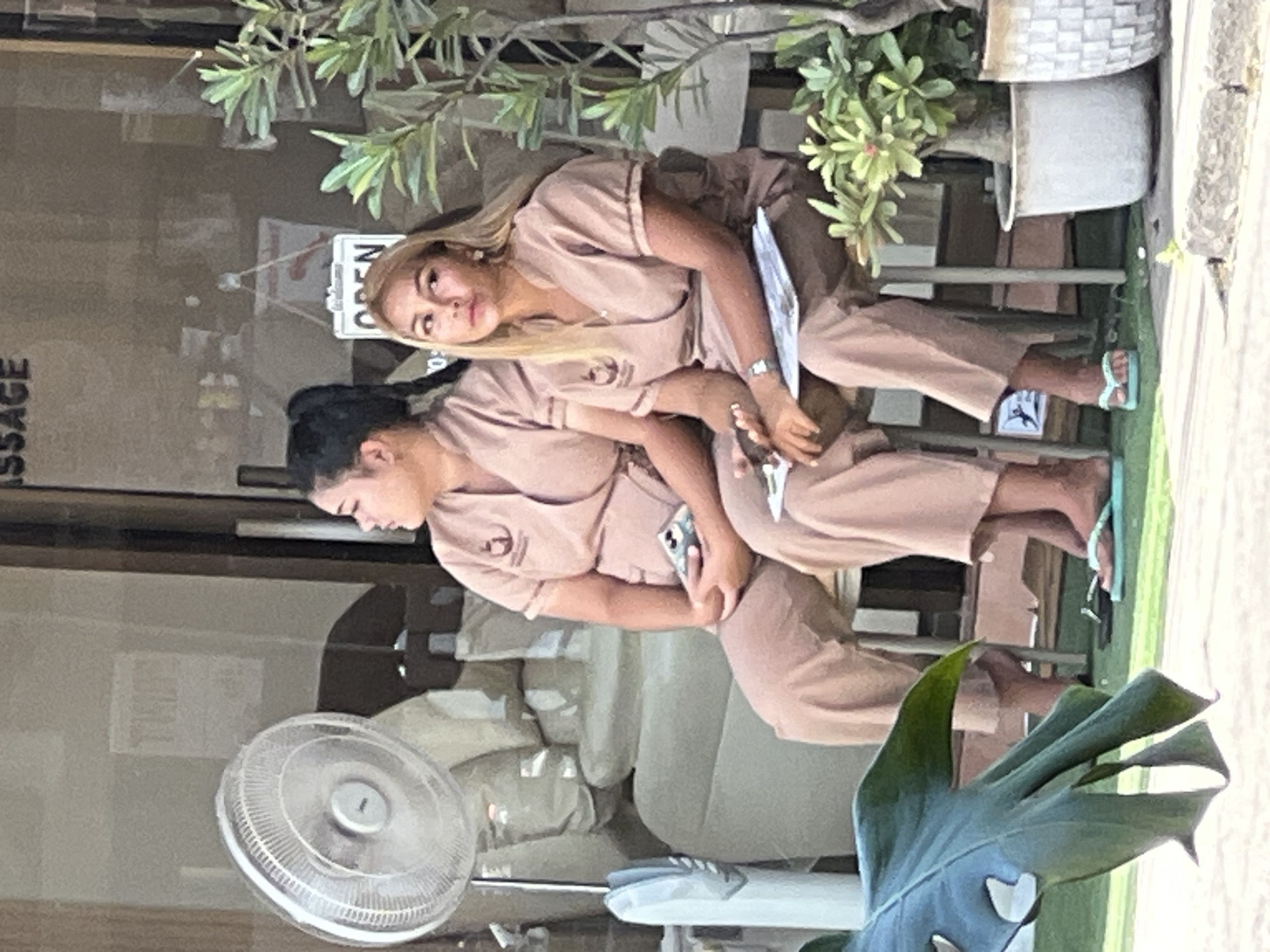 The image captures two women in matching tan uniforms seated outdoors, presumably in front of a business in Ko Samui District, Thailand. A large, white oscillating fan is visible to the left of the frame, positioned behind the women. The woman on the left has her dark hair pulled back in a ponytail and is looking to her left. She is holding a mobile phone in her lap. The woman on the right has blonde hair and is looking forward and slightly to her left. She is holding a clipboard with papers and a pen. Both women appear to be waiting. A leafy green plant is in a pot to the right of the frame. The background shows a window with text that includes the word "MASSAGE", and a white sign that says "OPEN". The ground is covered in a green matting, suggesting an outdoor seating area.