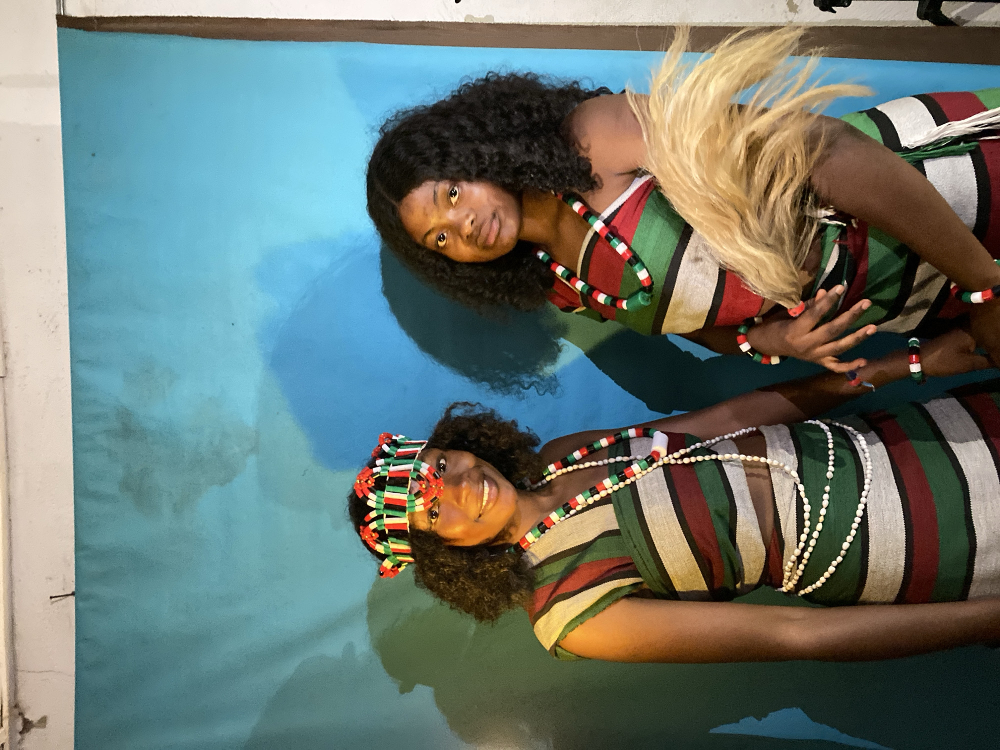 Two women are pictured in front of a blue backdrop. The woman on the top has dark curly hair, a necklace of black, red, green and white beads, and a dress with a striped pattern of white, red, and green. A plume of tan feathers is on her right side. She is wearing bracelets that match the necklace. The woman on the bottom has dark curly hair, a headdress of black, red, green and white, a necklace of white beads, and a dress with a striped pattern of white, red, and green. The location is unknown.
