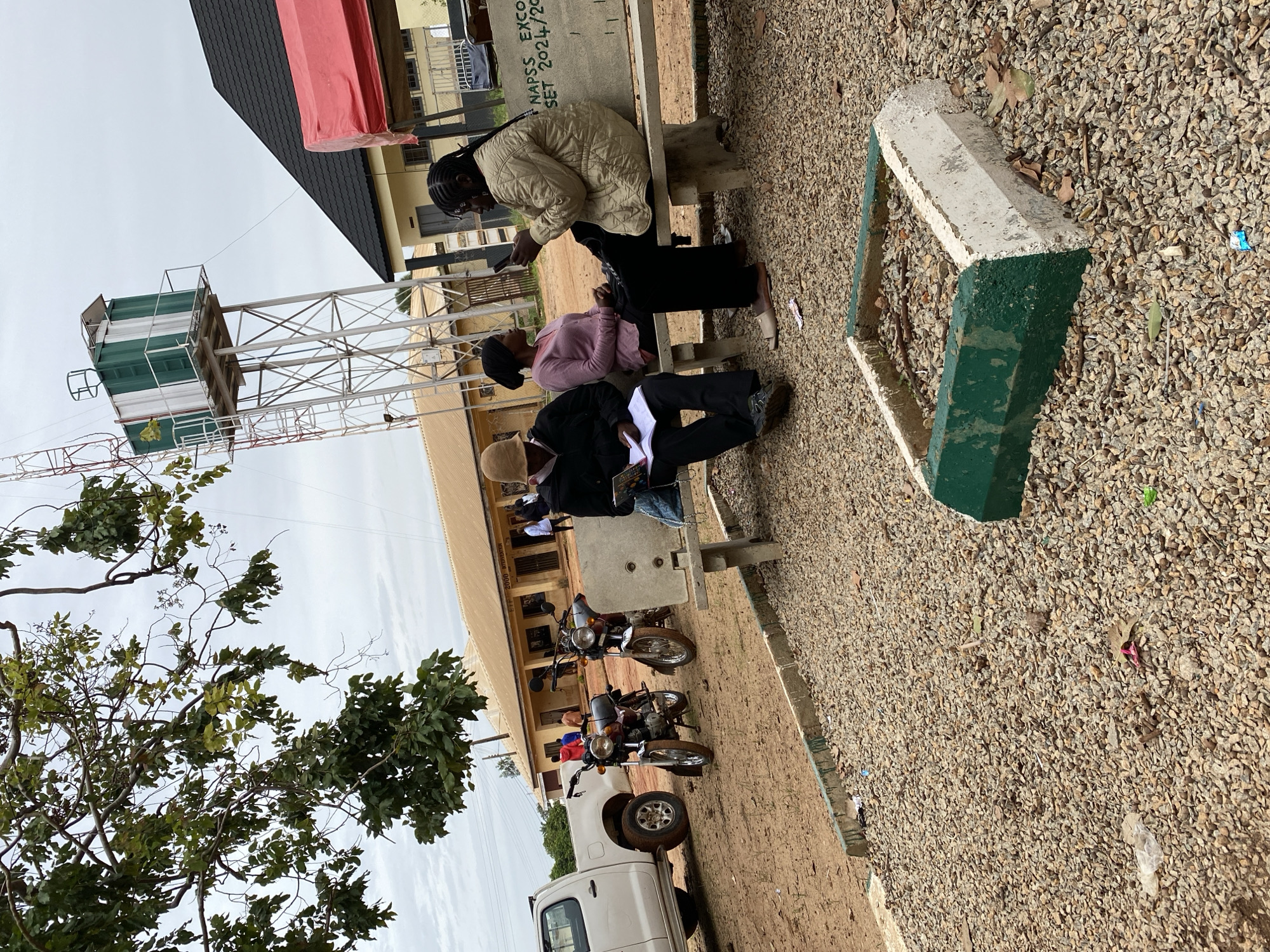 In Jos, Nigeria, this outdoor scene features a group of individuals gathered near a building. Three people are seated on a concrete bench. One person, wearing a light brown hat and dark jacket, is looking down at an open book or papers. Another person, in a pink sweater and black headscarf, is looking towards the person with the book. A third person, with braided hair and wearing a light beige jacket, is looking at their mobile phone. Behind them, a sign on the bench reads "NAPSS EXCO SET 2024/20".
In the background, several motorcycles are parked on the dusty ground. A white pickup truck is partially visible on the left side of the frame. The building appears to be a single-story structure with a brown roof and light-colored walls. A tall metal water tower with a green and white tank stands prominently to the left of the building. Trees with green leaves are present in the upper left portion of the image. The ground is covered in gravel and dirt. The sky is overcast.