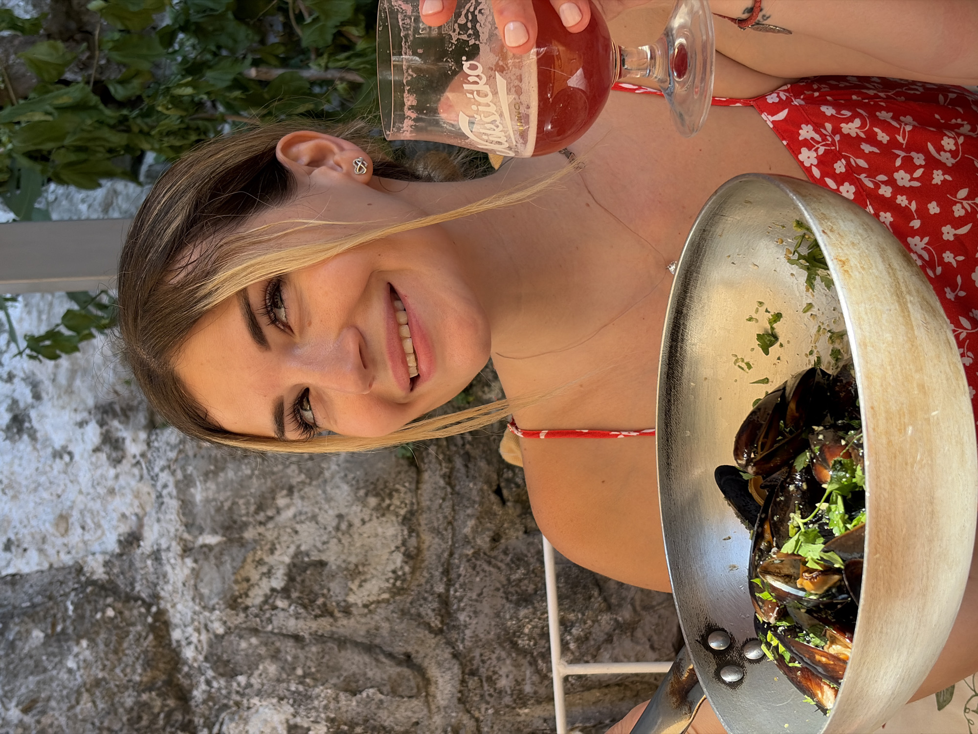 A young woman with blonde hair pulled back and strands framing her face is seated outdoors, holding a metal pan of mussels in her left arm and a glass of red liquid in her right hand. She is smiling and looking to her left, with a stone wall and green foliage forming the background. The pan, likely a serving dish, contains cooked mussels garnished with chopped green herbs. The glass has a foam head and the logo "Nikšićko" is visible. She is wearing a red dress with a white floral pattern and a delicate necklace. A white railing is visible behind her left shoulder. The setting appears to be a restaurant in Stari Bar, Montenegro, suggested by the "Nikšićko" beer logo, a popular Montenegrin brand, and the rustic stone architecture.