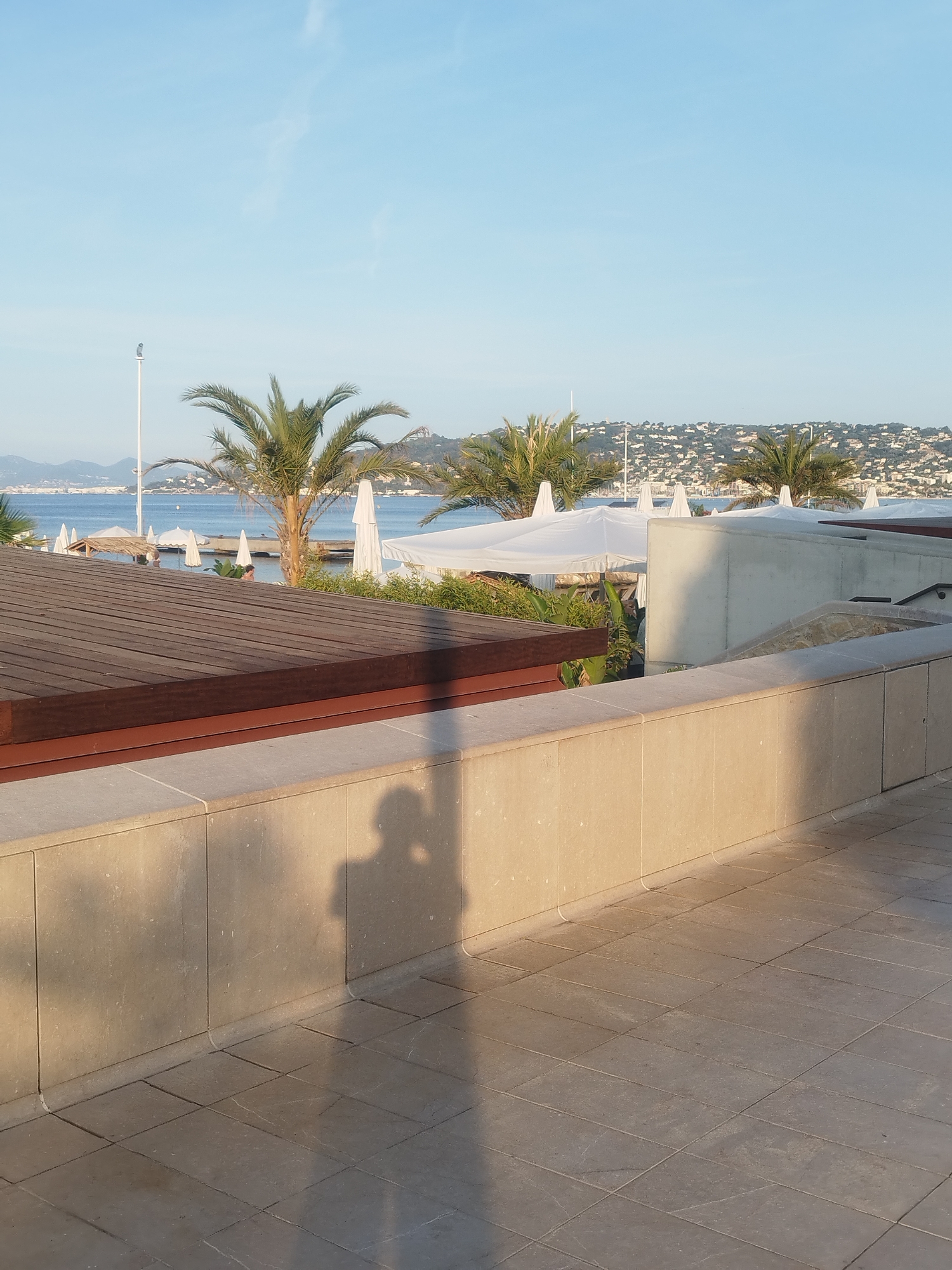 A sunny daytime outdoor scene in Antibes, France, captures a view of the Mediterranean Sea and a coastal town. In the foreground, a light-colored stone wall runs horizontally across the frame, with a tiled walkway extending from its base. A shadow of a person, possibly taking the photo, is cast on the wall and walkway. Beyond the wall, a wooden deck is visible, leading to a beach area with white umbrellas and palm trees. The sea stretches to the horizon, dotted with distant islands and a coastal town with white buildings nestled on a hill. The sky is a clear, pale blue.