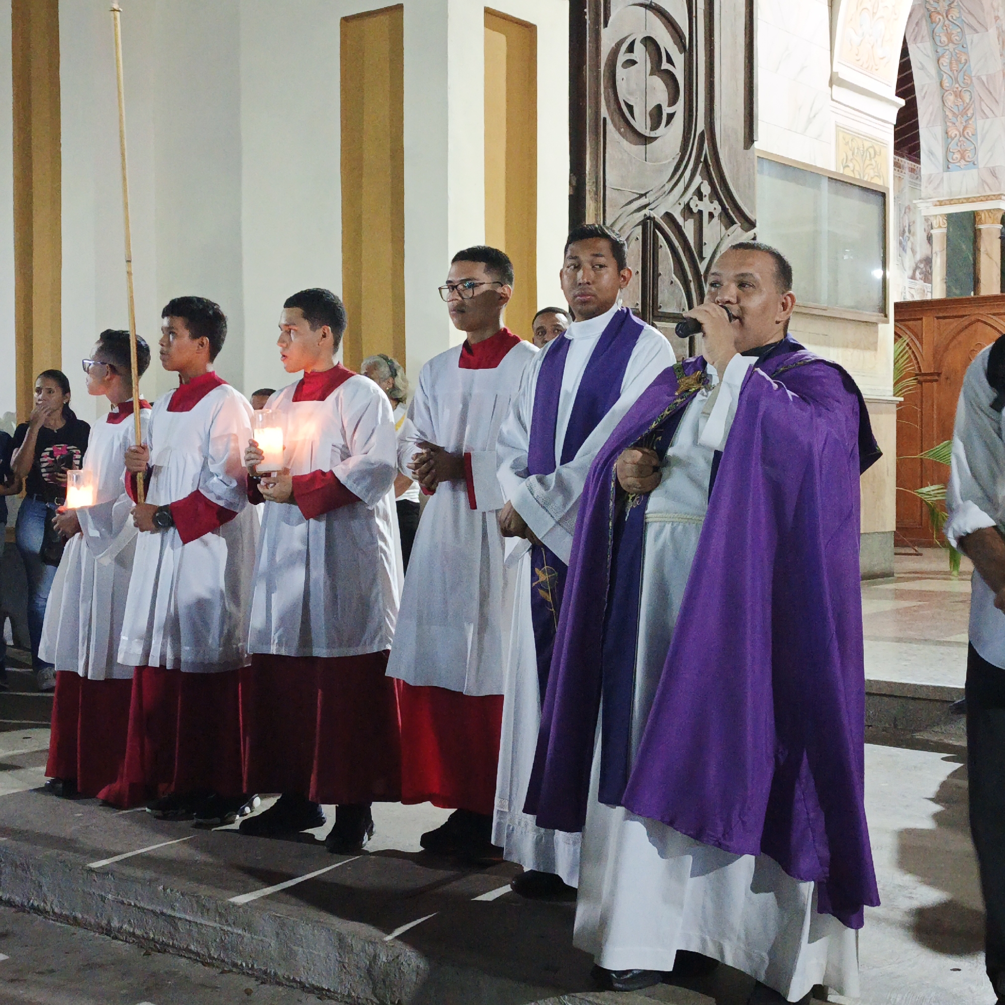 In Tinaquillo, Venezuela, a group of altar servers wearing white robes with red trim, one holding a tall bamboo rod, and one holding a lighted candle, stand on a stone platform alongside a priest who is speaking into a microphone. The priest is wearing a white alb, a purple stole, and a purple cope. They are in front of a large wooden door with decorative carvings. A few observers are in the background. The room is light, with columns in the background.
