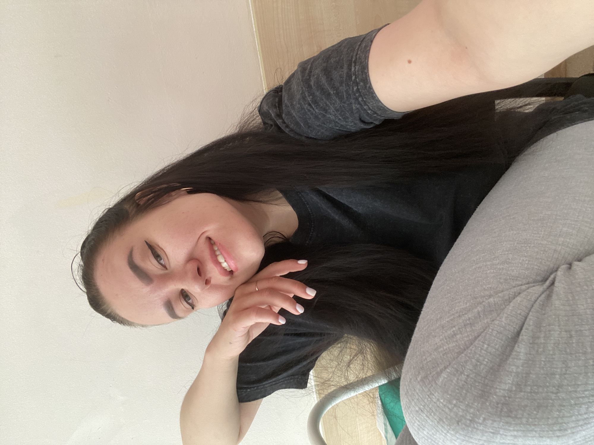 A selfie of a woman with long, dark hair and a black t-shirt. She is wearing grey sweatpants. The background consists of a white wall and a light wooden surface. The location is unknown.