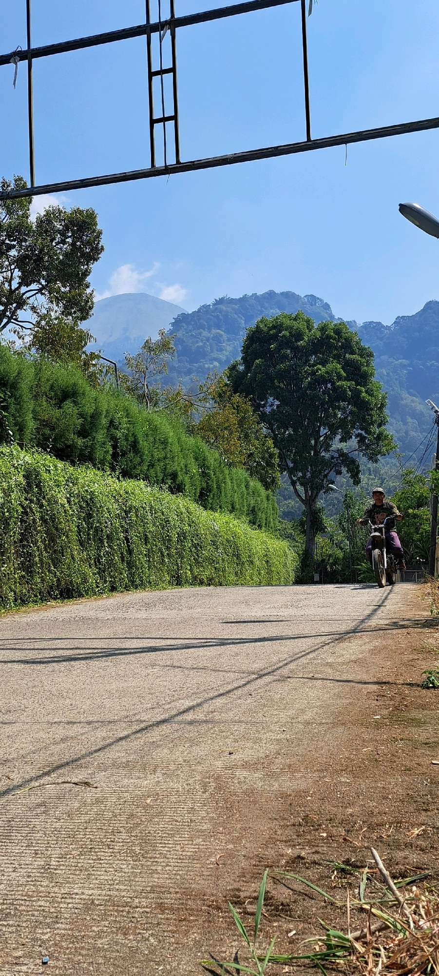 In the city of Duyung, Indonesia, a person is riding a motorcycle on a paved road. The road is bordered by lush green vegetation on the left and right. In the background, rolling hills covered in trees rise up to a hazy mountain peak under a clear blue sky. A metal structure is visible in the foreground, spanning across the top of the image. The scene is illuminated by bright sunlight, casting shadows across the road.