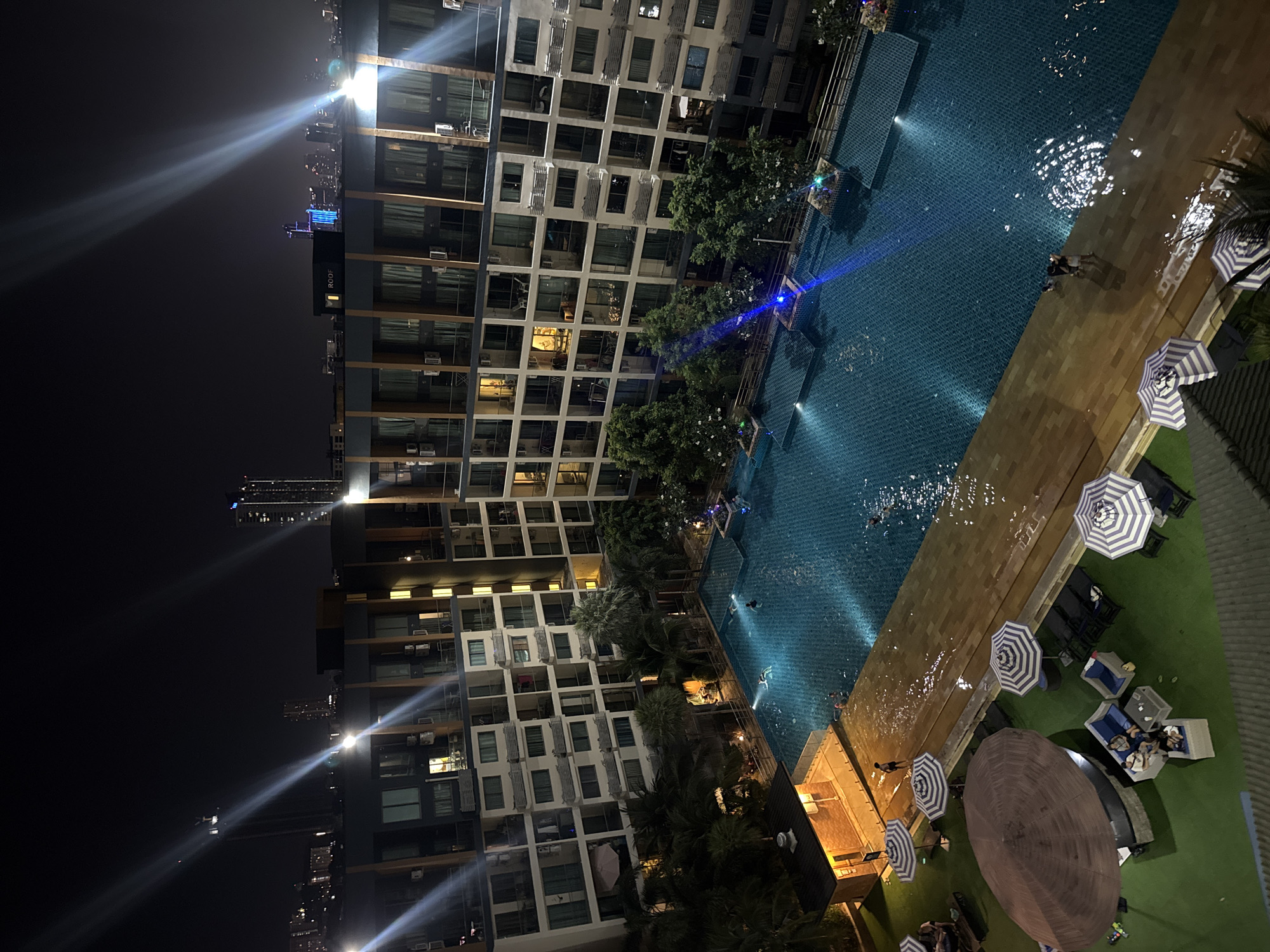 This nighttime photograph, taken in Pattaya City, Thailand, captures a vibrant scene from an elevated perspective overlooking a modern high-rise building, a swimming pool, and a recreational lounge area.
On the left, a multi-story building dominates the frame, featuring numerous rectangular windows and balconies. Many windows are illuminated, revealing interior lights, and air conditioning units are visible on the exterior walls. Beyond this building, the dark night sky is dotted with distant city lights, some producing prominent lens flares. On one of these distant high-rises, the word "ROOF" is visible on a sign.
In the mid-ground, a large, blue-tiled swimming pool is brightly lit by underwater lights, casting an inviting glow and creating ripples on its surface. Several people are visible in the pool, swimming or standing in the water. Adjacent to the pool, a section of light-brown decking provides a transition to the lounge area.
To the right, a verdant artificial turf area is set up with various outdoor furniture. This includes several lounge chairs, multiple blue and white striped umbrellas, and one large, circular brown umbrella. A few individuals are seen relaxing on outdoor sofas and chairs in this area. Palm trees and other foliage are interspersed between the building and the pool, adding to the resort-like atmosphere. The overall scene conveys a sense of nighttime leisure and activity.