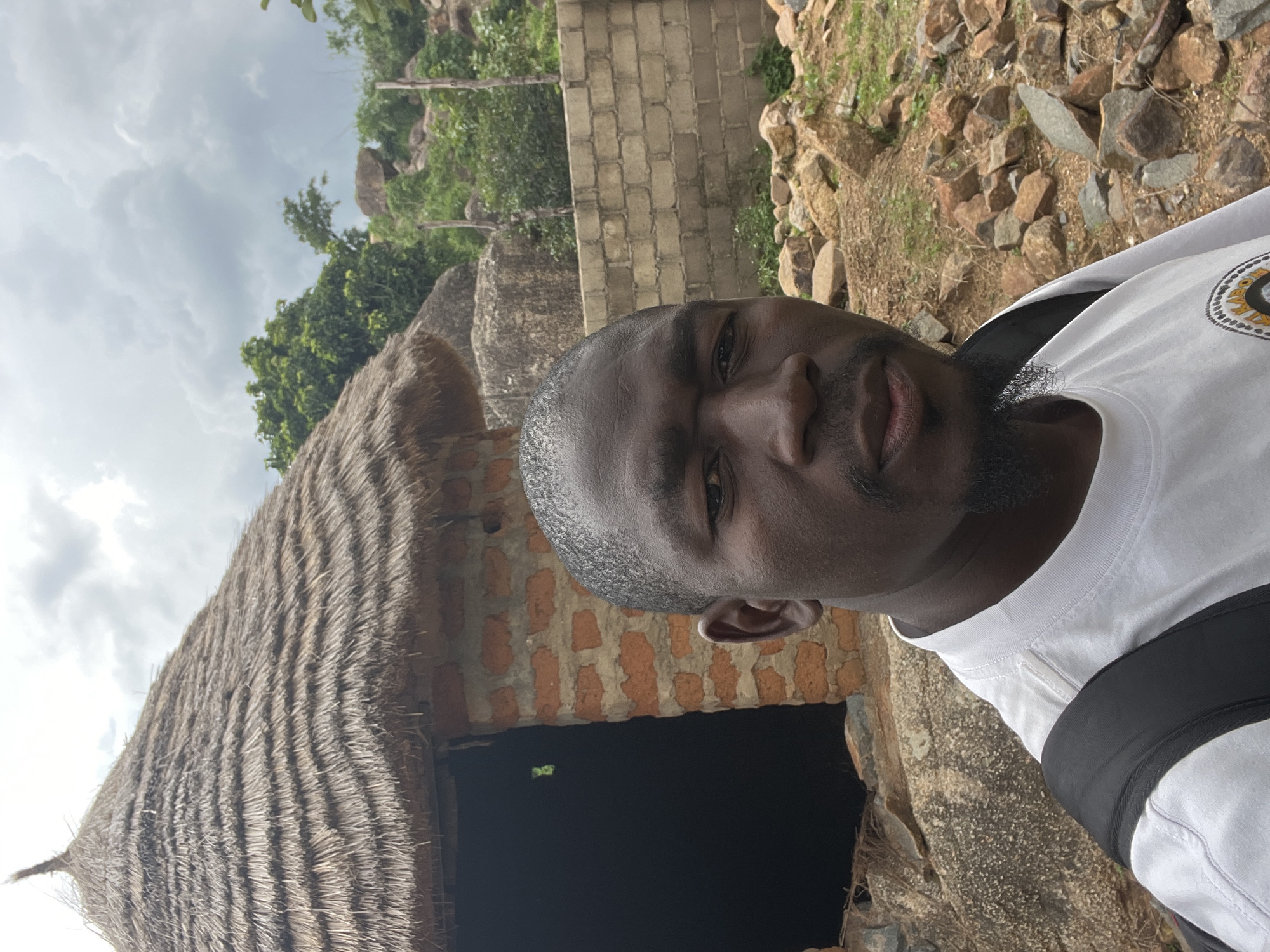 A dark-skinned man with a goatee and short hair is taking a selfie outdoors. He is wearing a white t-shirt with a black backpack strap visible over his left shoulder. Behind him is a small, round building with a thatched roof made of straw. The walls of the building appear to be constructed from red bricks. To the right of the building, there is a low wall made of concrete blocks. The background consists of a rocky, hilly landscape with green vegetation and trees, under a cloudy sky. The ground in front of the man is covered with loose rocks and dirt. The image appears to be taken in a rural or semi-rural setting in Jos, Nigeria.