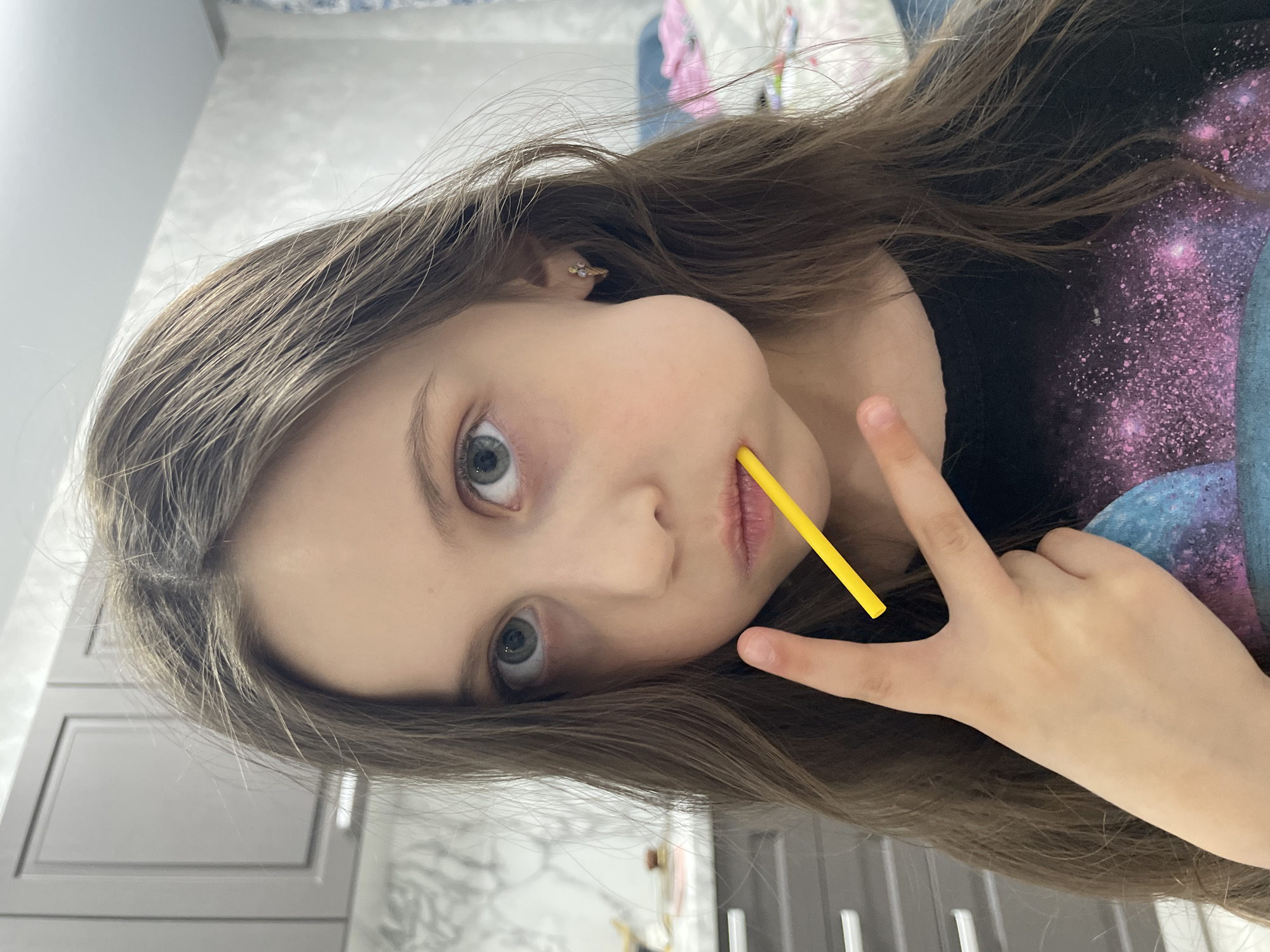 A young girl is seen close-up, holding a yellow straw near her mouth and making a peace sign with her hand. She has long brown hair, fair skin, and blue eyes. She is wearing a dark-colored t-shirt with a galaxy print. The background appears to be an interior setting with a white wall and gray cabinets. The location in Izhevsk, Russia is not evident from the image.