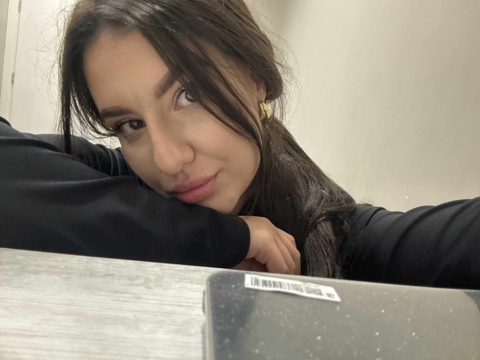 A young woman with dark hair is resting her head and arm on a desk or table with a gray, wood-grain pattern. She is wearing a black long-sleeved shirt or sweater. Part of a rectangular object with a barcode is visible in the foreground. The background is a plain, light-colored wall. The setting appears to be indoors, possibly an office or home. There is no information available to determine the location in the city of Burgas, Bulgaria.