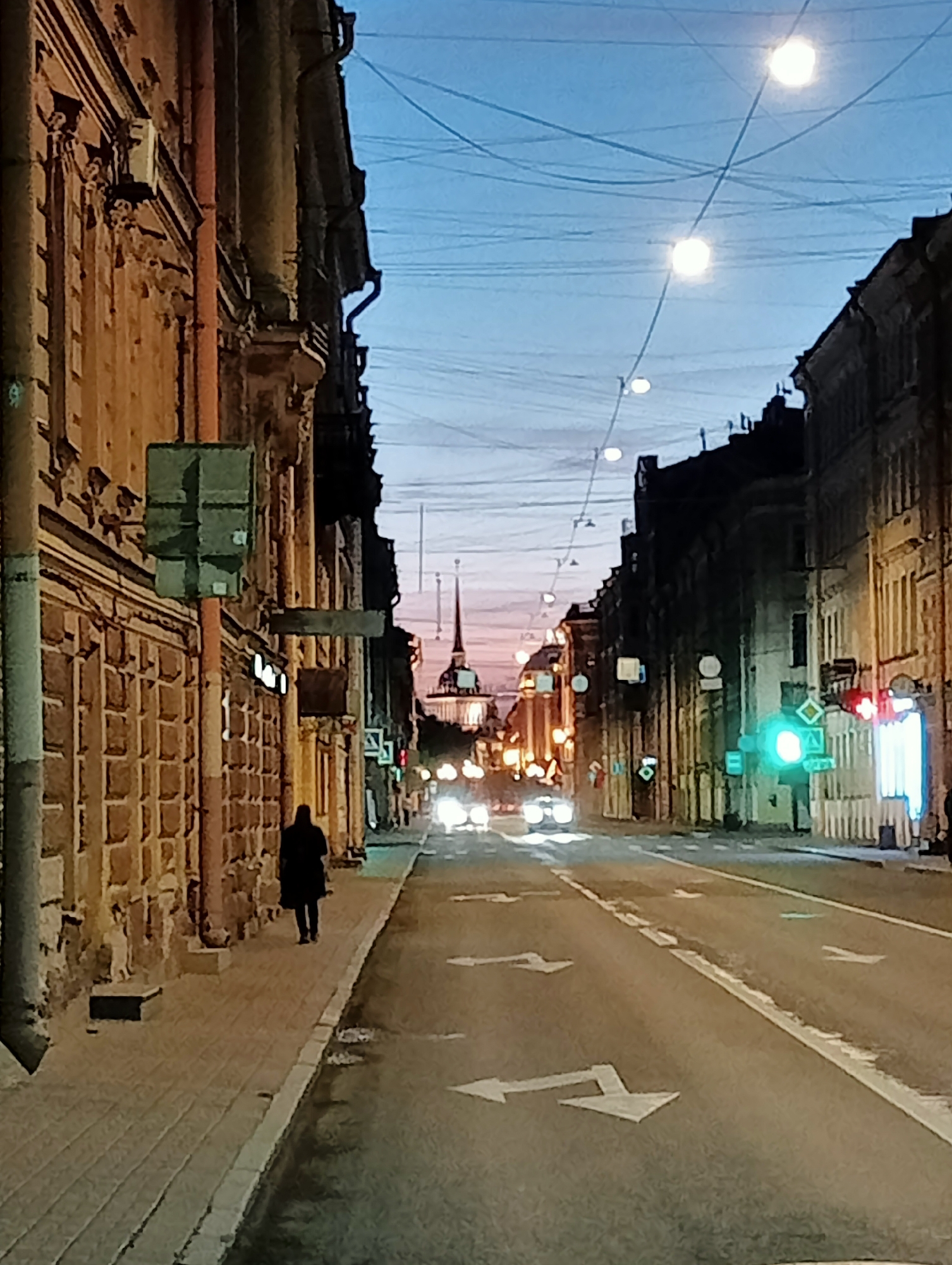 This image depicts a street scene in Saint Petersburg, Russia, likely during twilight or early evening. The perspective is from street level, looking down a relatively wide, asphalt road.
On the left side of the street, a building with an ornate, light-brown facade is visible. The building features architectural details like decorative cornices and window surrounds. A sidewalk runs alongside the building, paved with rectangular stones. A lone figure, silhouetted and dressed in dark clothing, is walking away from the viewer on the sidewalk. Several traffic signs are affixed to poles along this side of the street, including a green rectangular sign and a gray rectangular sign. A reddish pipe runs vertically down the side of the building.
On the right side of the street, another building with a darker facade rises up, its details less distinct. Several streetlights, emitting a bright white light, are suspended across the street by wires. The sky above is a gradient of pale blue and pinkish hues, indicating the time of day.
In the distance, the spire of a building, possibly a church or government structure, is visible against the sky. The road is marked with white directional arrows. Several cars are present on the road, their headlights illuminated, suggesting they are in motion. Traffic lights are visible, with one displaying a bright green light and another a red light. The road surface appears damp in places, with some reflections. The overall atmosphere is urban and somewhat tranquil, characteristic of a European city at dusk.