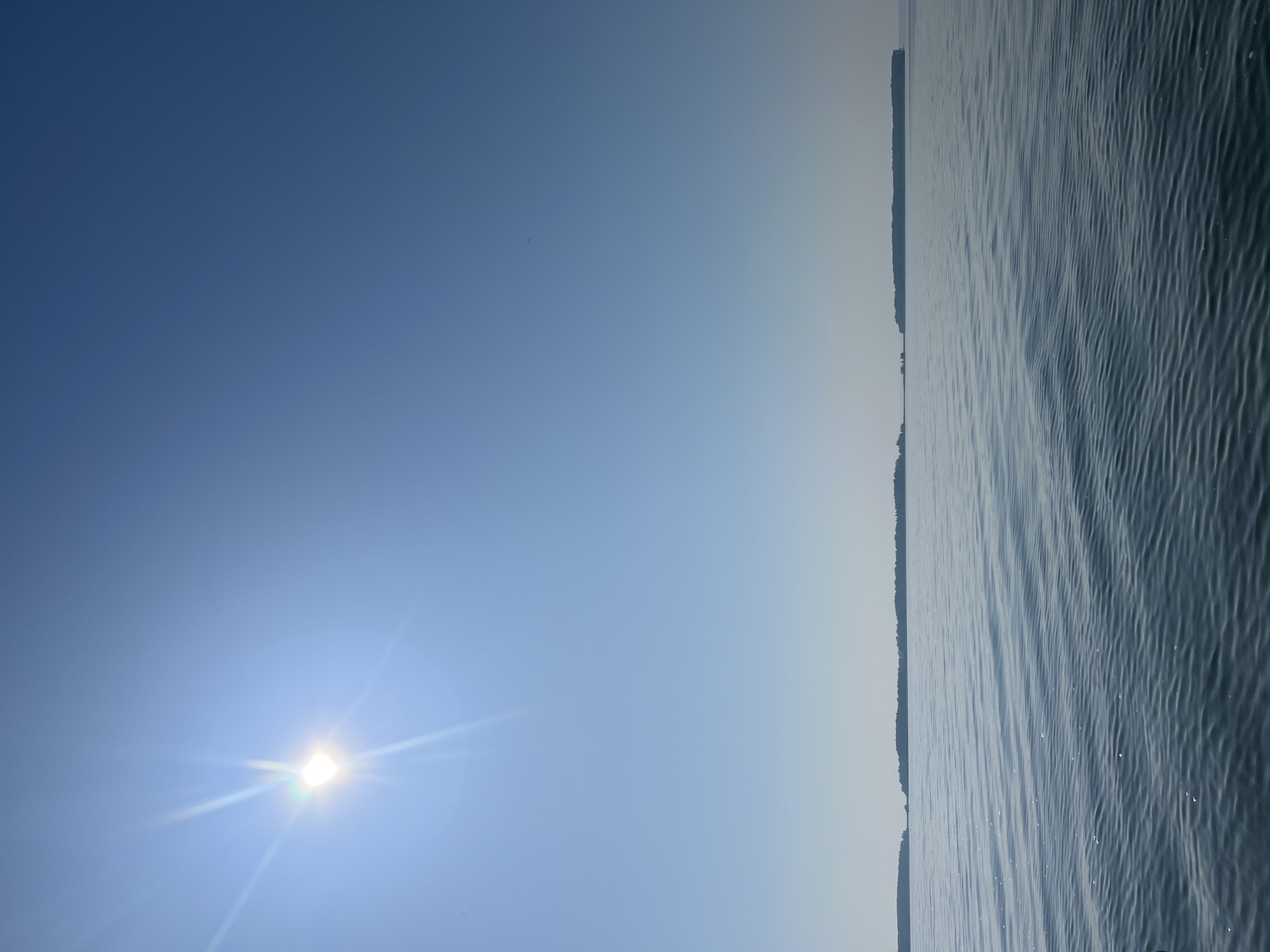 This is a vertical, eye-level shot of a body of water under a bright blue sky. The sun is visible in the upper left corner, casting a bright glare and lens flares that extend across the upper portion of the image. In the distance, a series of low, tree-covered islands or landmasses stretch horizontally across the horizon, dividing the water from the sky. The water in the foreground is calm with gentle ripples, reflecting the sunlight and creating a textured surface. The scene is devoid of any people or specific man-made structures, evoking a sense of natural tranquility. The image is believed to have been taken in the vicinity of Novopichugovo, Russia, a location known for its natural beauty and bodies of water.