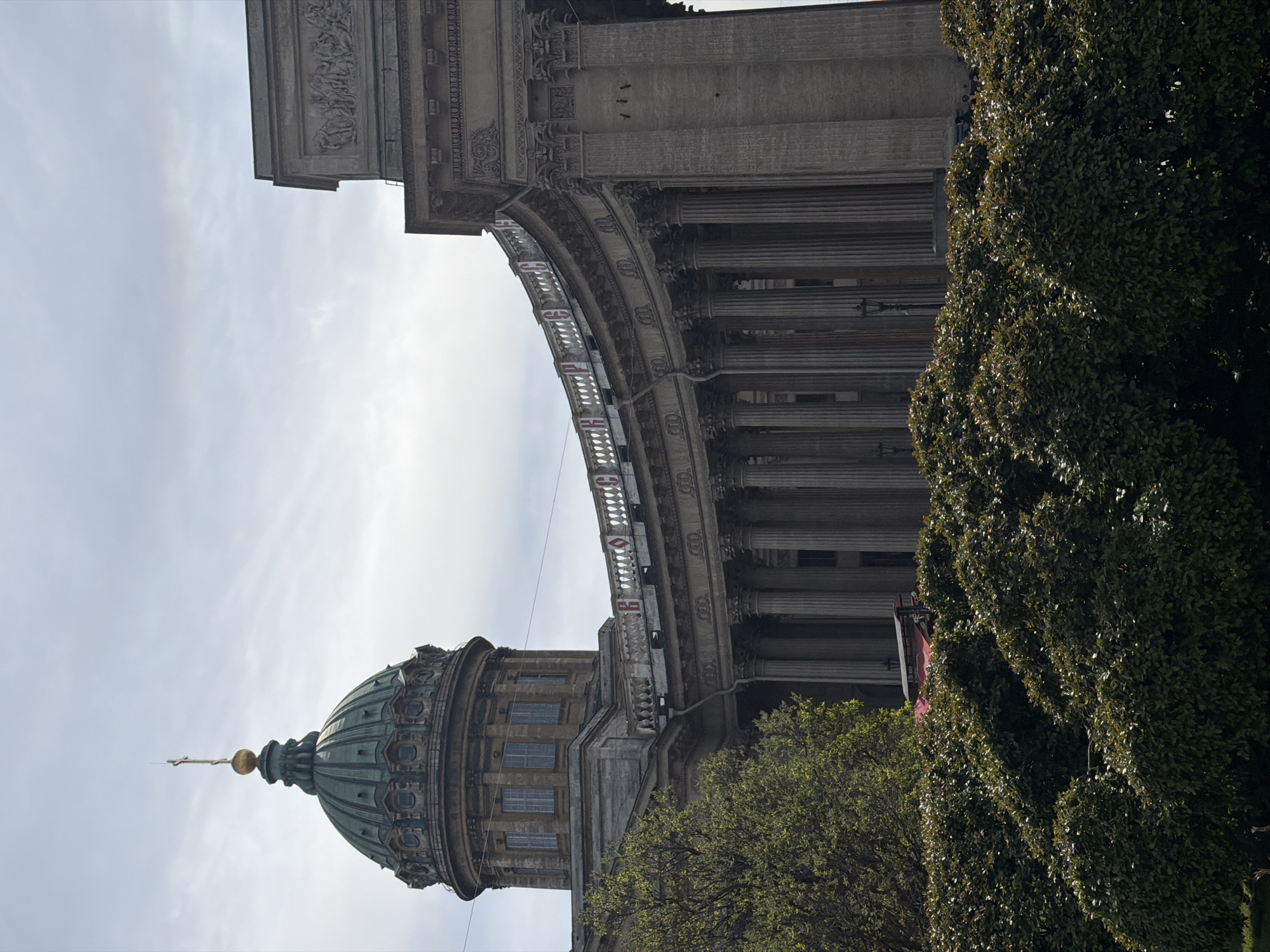 The image shows a partial view of the Kazan Cathedral in Saint Petersburg, Russia, partially obscured by trees on the right. The perspective is from the ground looking up, with the image rotated to the side. The iconic green dome of the cathedral is visible on the left, along with its facade and windows. A portion of the cathedral's colonnade and decorative elements are seen stretching upwards. The sky is visible in the background.