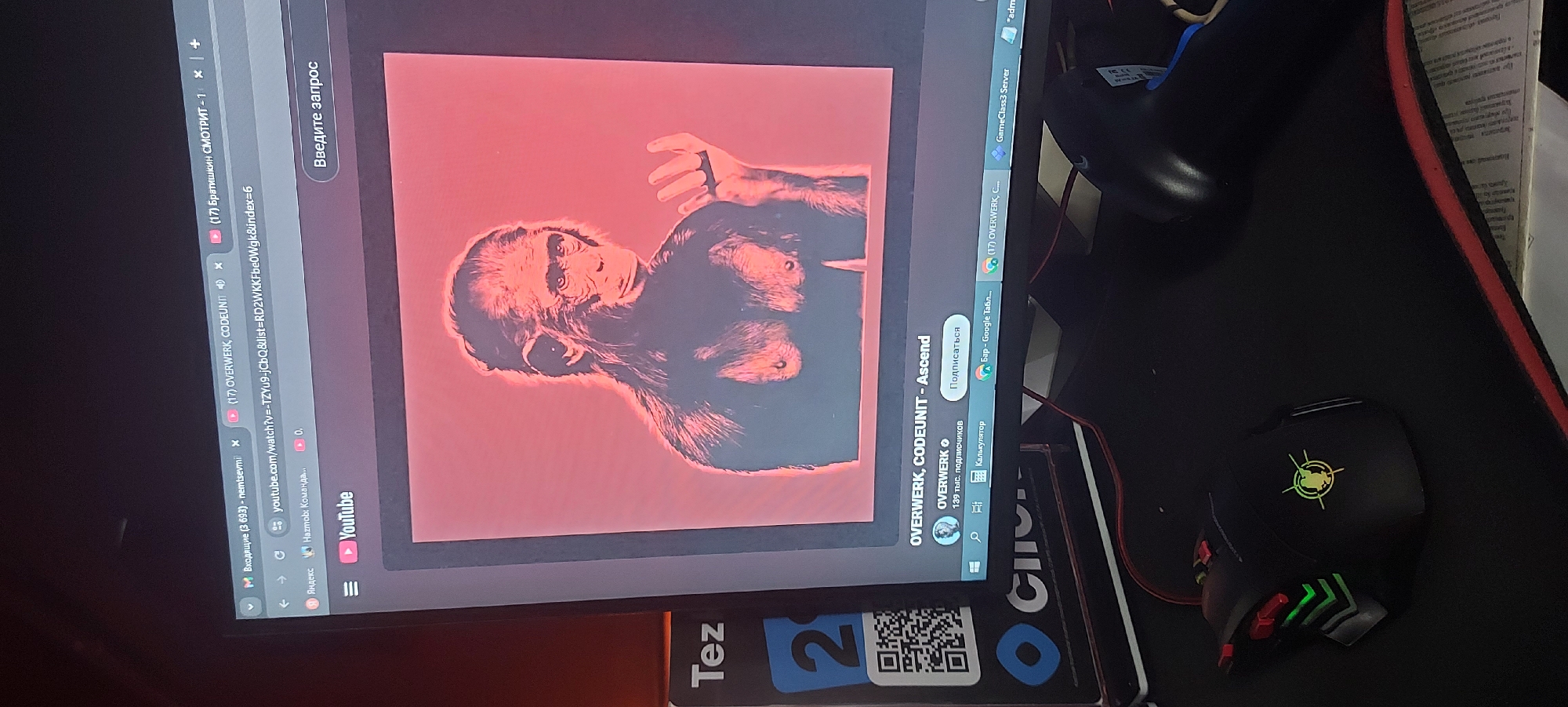 The image shows a computer screen displaying a YouTube video of "OVERWERK, CODEUNIT - Ascend," featuring an image of a chimpanzee. The web browser also has tabs open for "Входящие (3 693) - nemtsevmi" and "(17) Братишкин СМОТРИТ-1." The browser address bar shows the YouTube URL. On the desktop taskbar, there are also opened applications like "GameClass3 Server" and "Бар - Google Табл..."
Next to the computer, on a desk, are a gaming mouse, a partially visible keyboard, and some papers. There is a card with the word "Tez" and a QR code visible beneath the monitor. There is nothing in the image to definitively indicate the location is Tashkent, Uzbekistan.