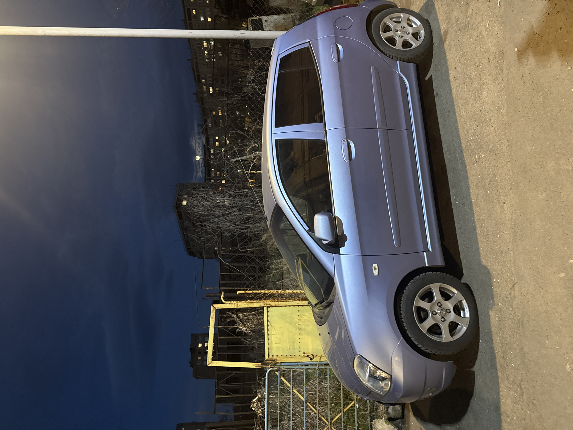 The image depicts a light blue Kia Picanto parked on a concrete surface at night in what appears to be Yerevan, Armenia. In the background, there are multi-story residential buildings with lit windows, suggesting it's evening. There's a thin white pole visible in the upper left. A fence is present near the car, with a yellow and rusted metal structure behind it. The sky is dark, with a moon visible between the buildings.