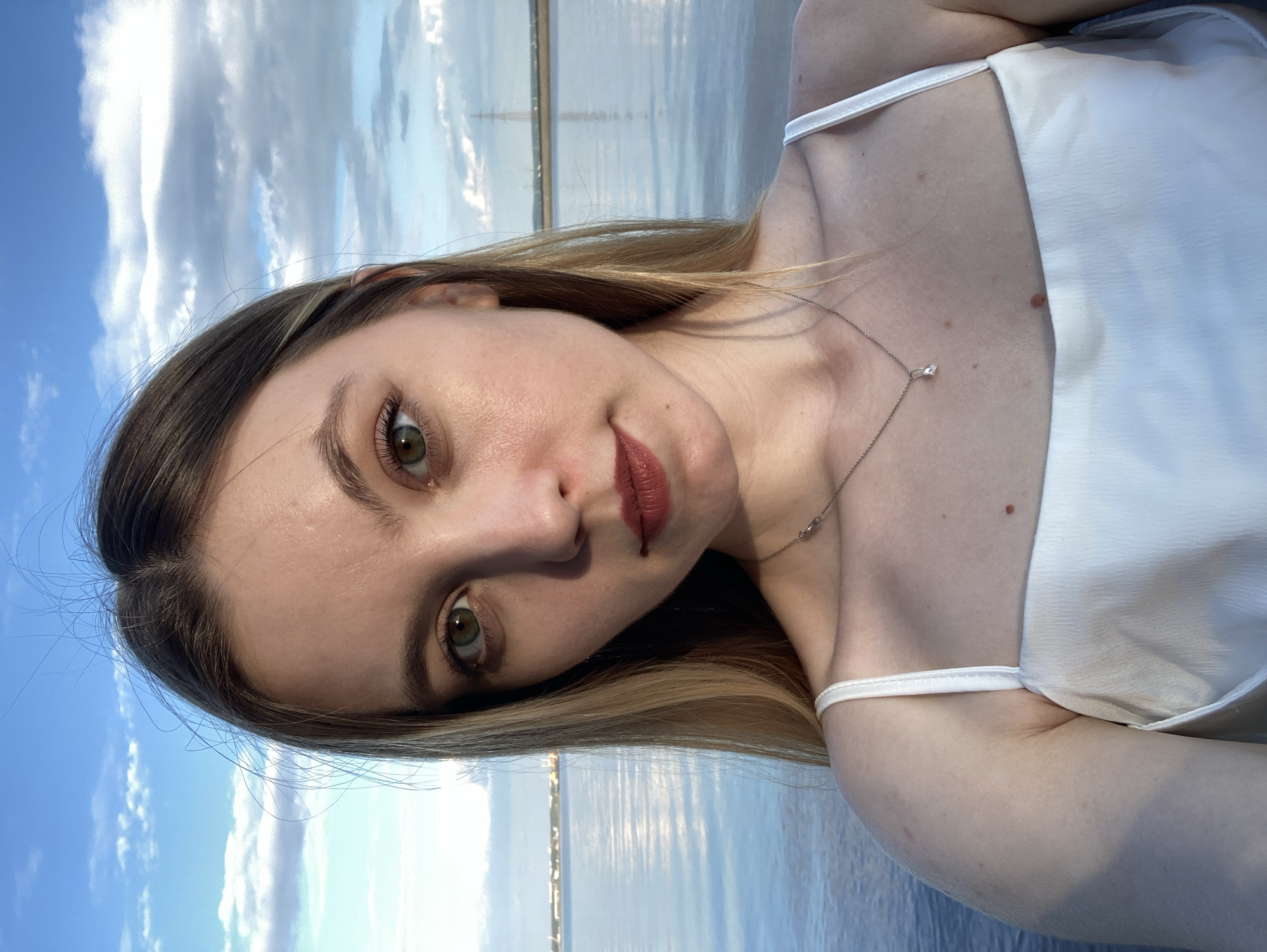 A young woman with blonde hair and green eyes is pictured from the chest up, taking a selfie outdoors near a body of water. She is wearing a white satin camisole and a delicate silver necklace with a small pendant. Her makeup includes mascara, eyeliner, and a matte reddish-brown lipstick. The background consists of a calm body of water with reflections of the sky, which is blue with scattered white clouds. The horizon line shows a distant shoreline with some structures and a power line tower. The overall lighting suggests it is late afternoon or early evening. The location appears to be Noviy, Russia, given the context provided.