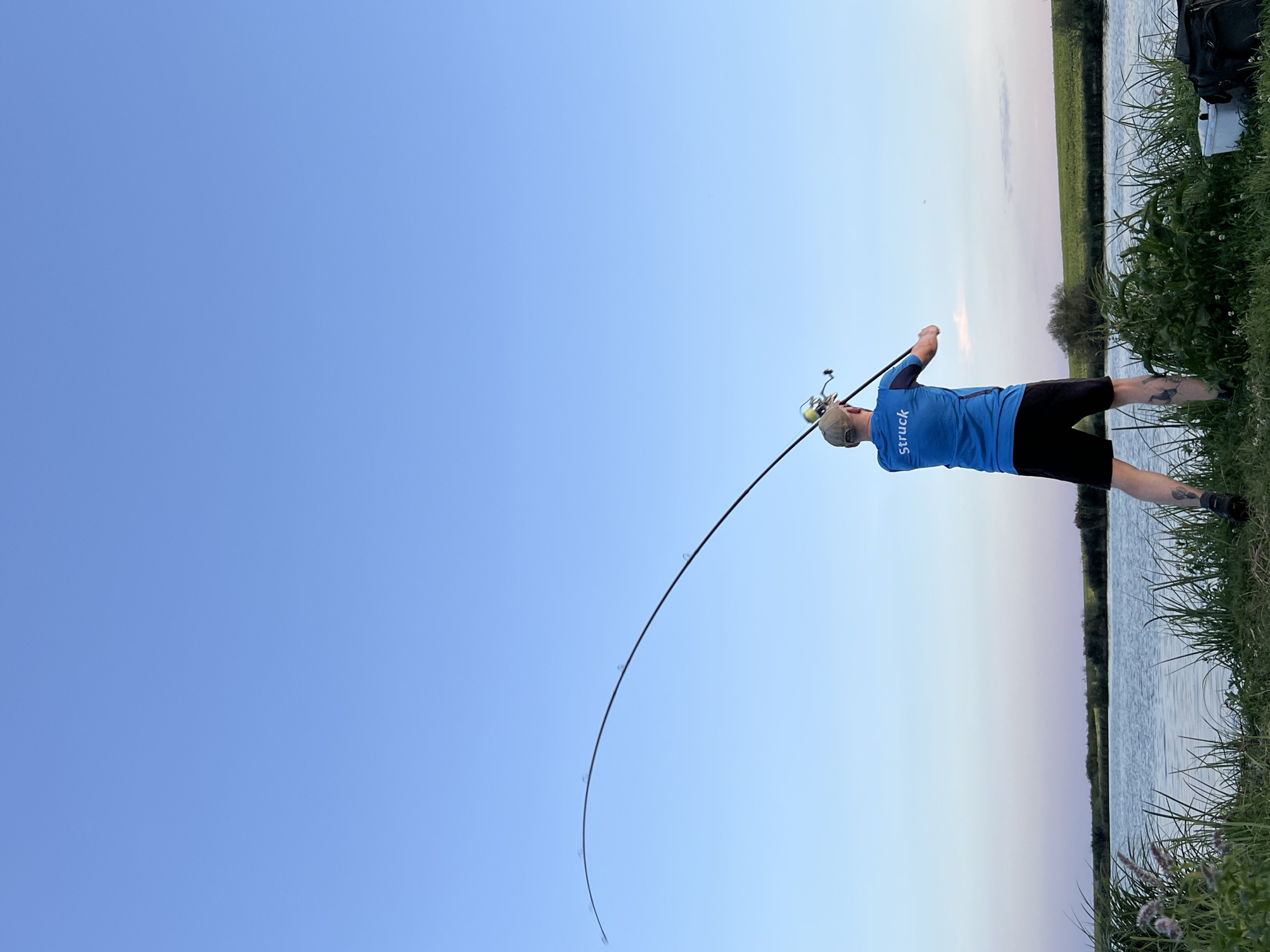 A man is fishing on the bank of a body of water, likely a river or lake, in Mateushivka, Ukraine. The sky is a clear, pale blue with a hint of pink near the horizon, suggesting it is either early morning or late evening. The man is wearing a blue t-shirt with "Struck" written on the back in white, black shorts, and a baseball cap. He is holding a fishing rod, which is bent considerably, indicating he has likely caught something. He is positioned facing away from the camera, towards the water. In the foreground, tall green grass and some lighter-colored reeds are visible. To the right, a white bucket and a dark bag are placed on the ground. The water is calm, reflecting the sky. In the background, there is a low embankment covered in green vegetation, with some trees further in the distance. The overall scene depicts a tranquil moment of outdoor activity.
