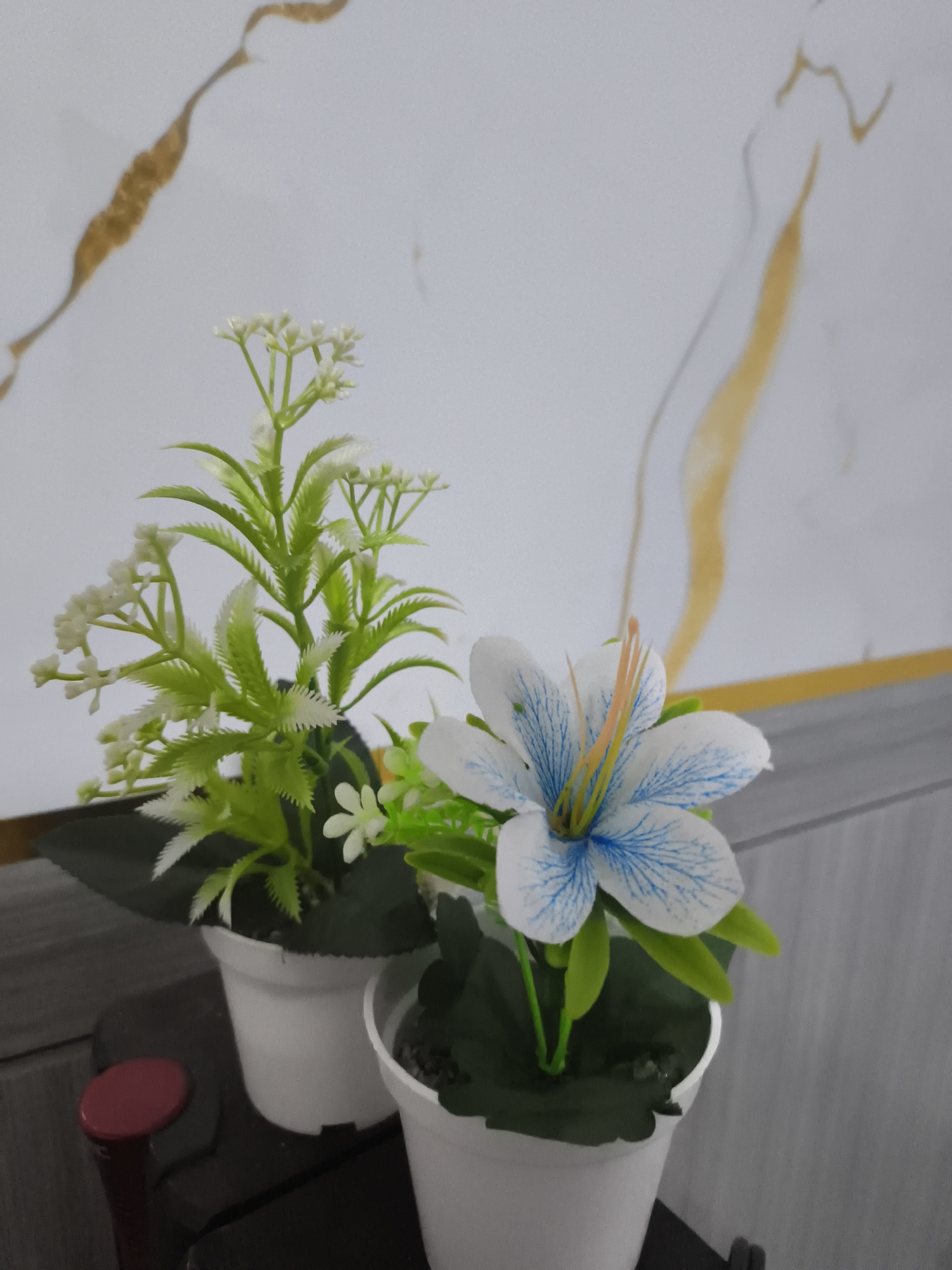 Two artificial potted plants are displayed in front of a white wall with abstract golden and grey streaks. The plant on the left features green foliage and small white flowers. The plant on the right has a single white and blue artificial flower with delicate blue lines on its petals and yellow stamens. Both plants are in white plastic pots. The plants are placed on a dark surface, possibly a table or shelf, and a red cylindrical object is partially visible on the left. The lighting suggests an indoor setting, possibly a home or office in Pangkalpinang, Indonesia.