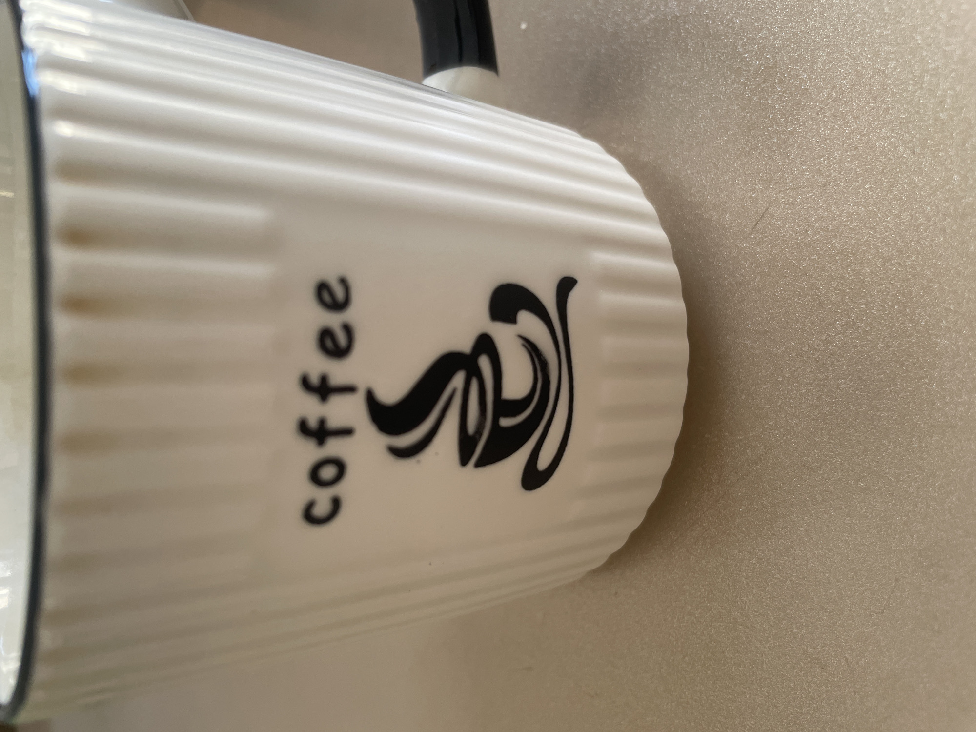 A close-up, slightly angled shot shows the upper portion of a white ceramic mug with vertical ridges. The word "coffee" is printed in black font above a stylized black graphic of a coffee cup with steam rising. The mug has a black handle that is partially visible on the right side. The mug sits on a light-colored, textured surface that appears to be a countertop. The lighting is even, and the background is blurred, suggesting the focus is solely on the mug. The image does not contain any information related to Tashkent, Uzbekistan, or any urban environment.