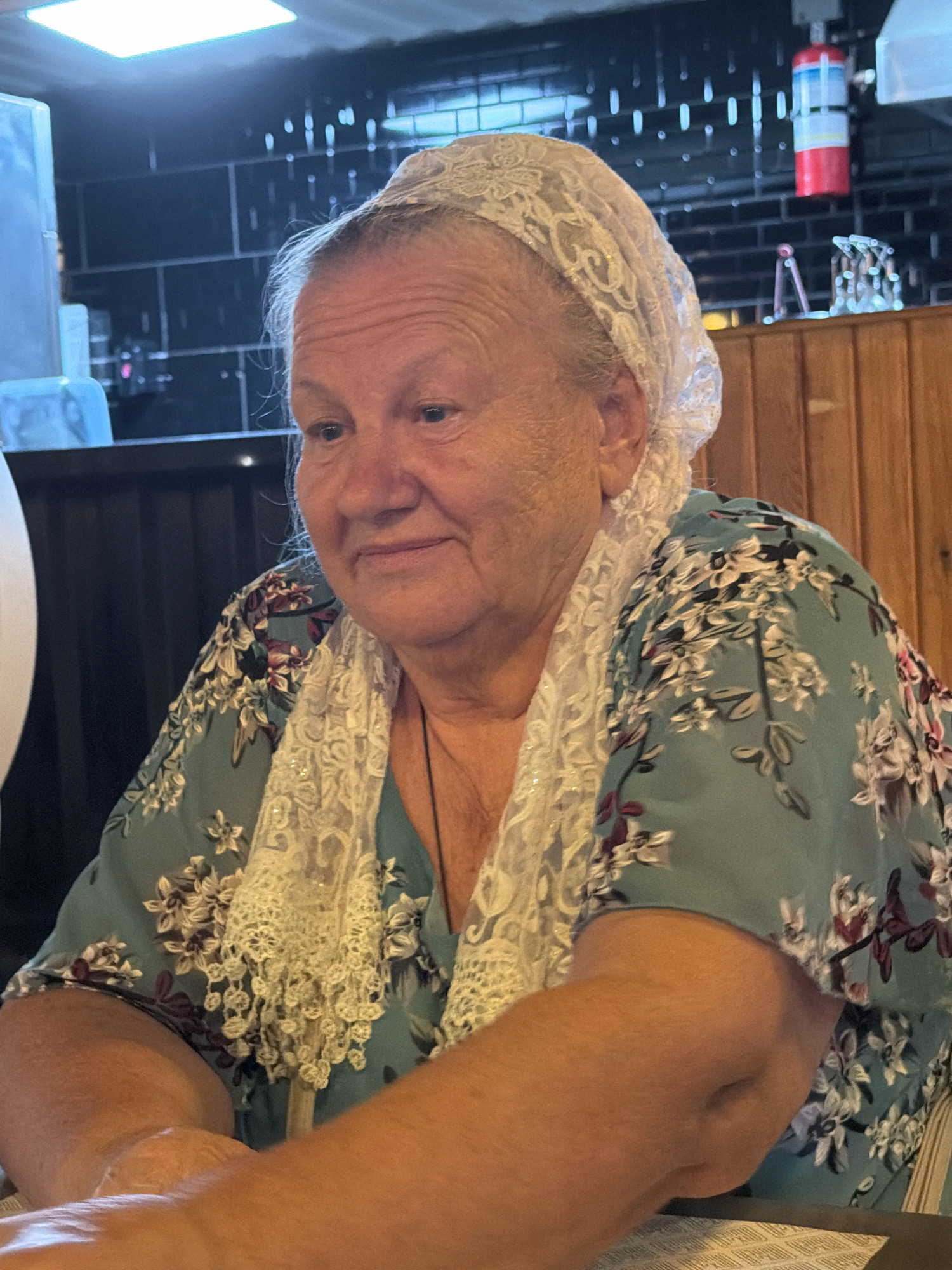 A medium close-up shot captures a woman with fair skin, light hair, and blue eyes, wearing a lace headscarf and a teal floral-patterned dress. She is seated at a table, her arms resting in front of her. The background includes dark, tiled walls, wooden paneling, and a red and white object hanging on the wall. The lighting suggests an indoor setting, possibly a restaurant. No specific location in Sharm El Sheikh, Egypt can be determined from the image.