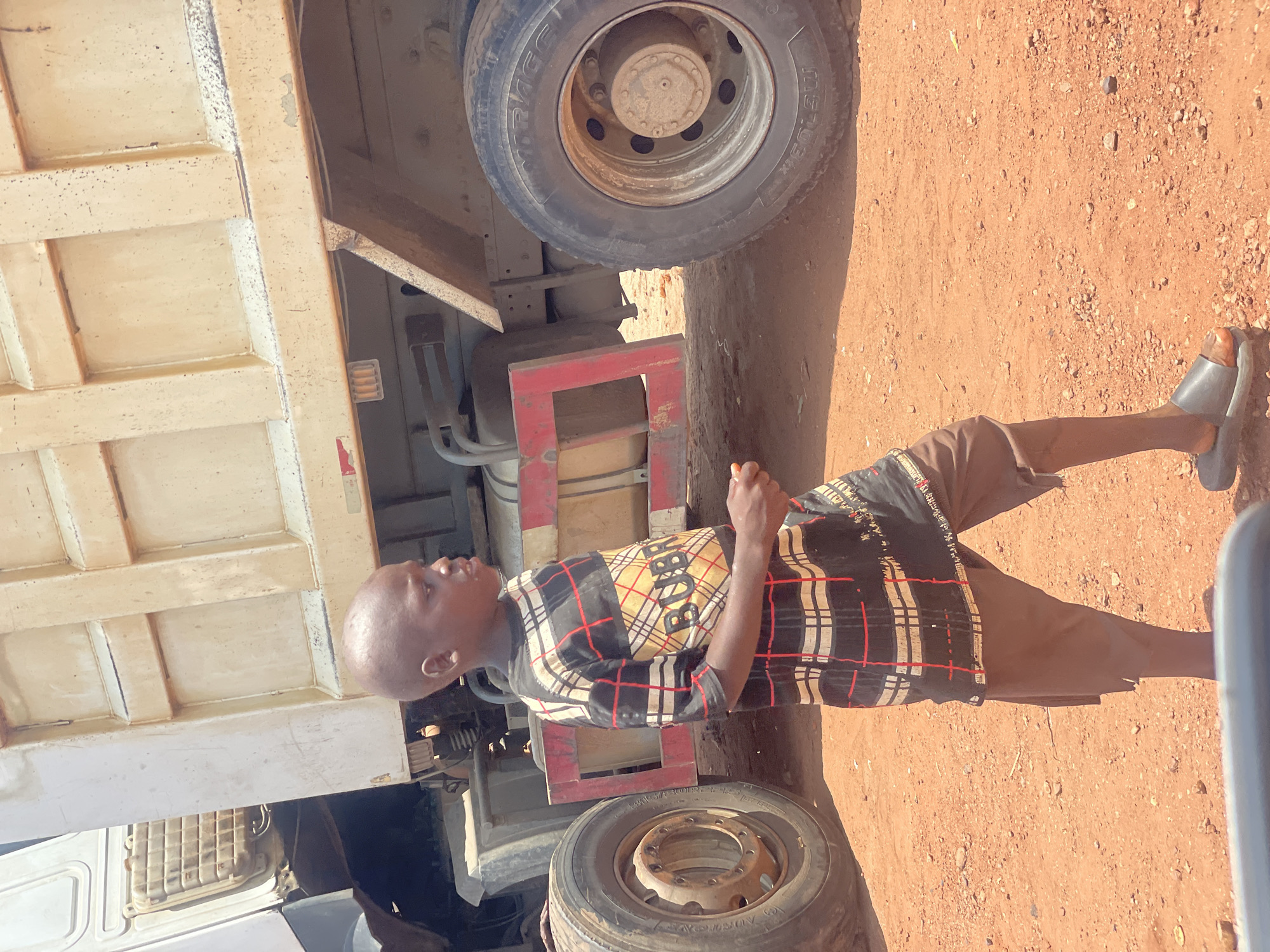 A person wearing brown shorts and a plaid shirt stands next to a large truck on a dirt surface in Kwali, Nigeria. The person wears black sandals on their feet. The truck has two visible tires, and a beige rectangular component is visible on the left side of the image. The surface is reddish-brown dirt with some small rocks or pebbles.