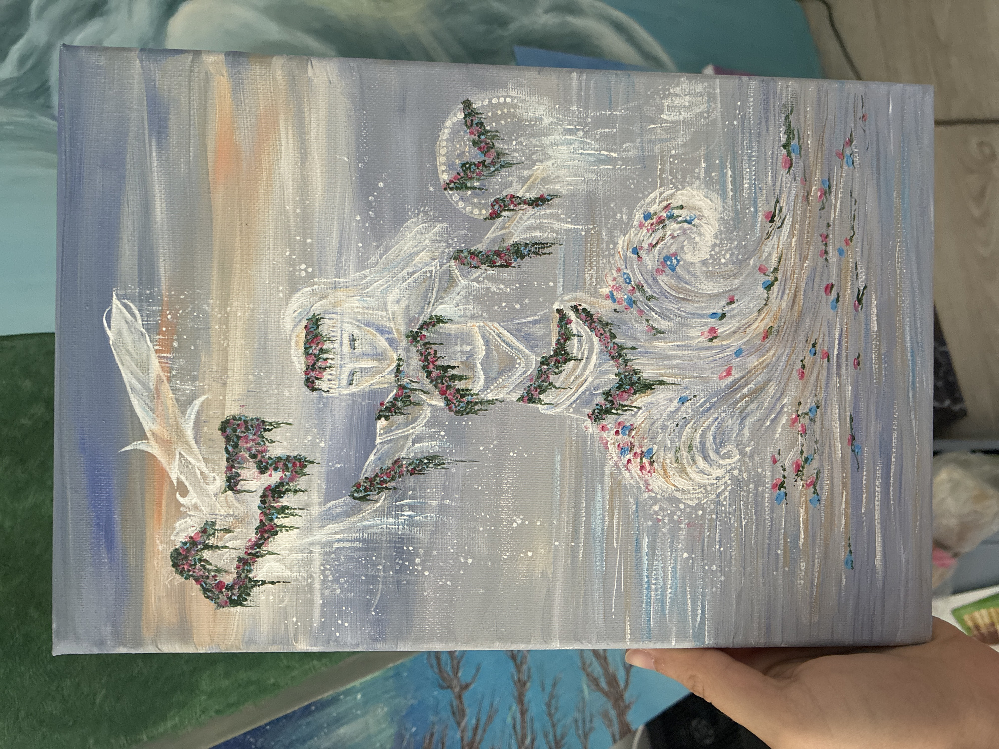 The image shows a hand holding a painted canvas. The painting depicts a white, abstract figure with floral accents on a background of blended blues, grays, and a touch of peach. The figure has a flowing, almost ethereal quality. The location is unspecified.